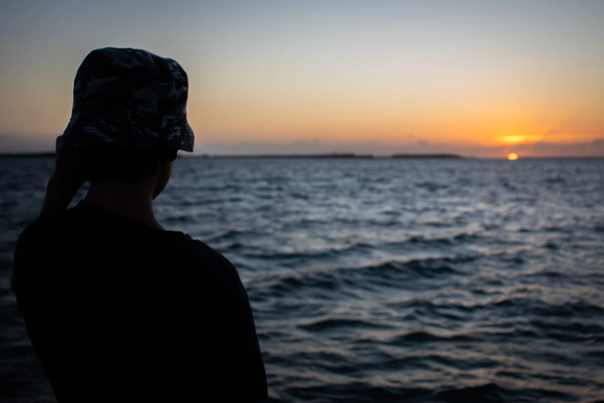 Person wearing a camouflage hat looking at a sunset over the ocean.