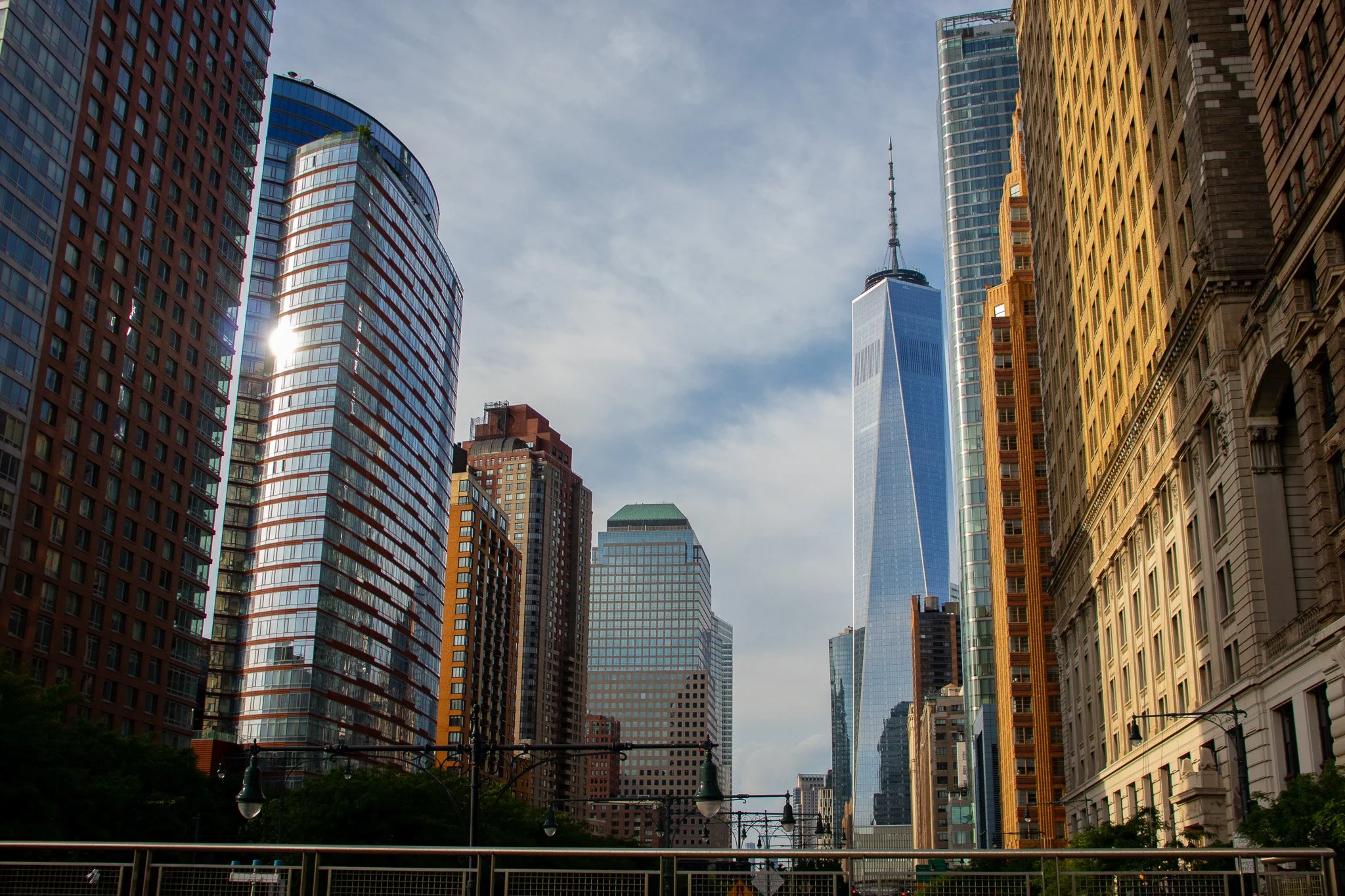 Cityscape with tall modern skyscrapers including One World Trade Center, some with glass facades reflecting sunlight, and older buildings with ornate architecture at street level, under a partly cloudy sky.