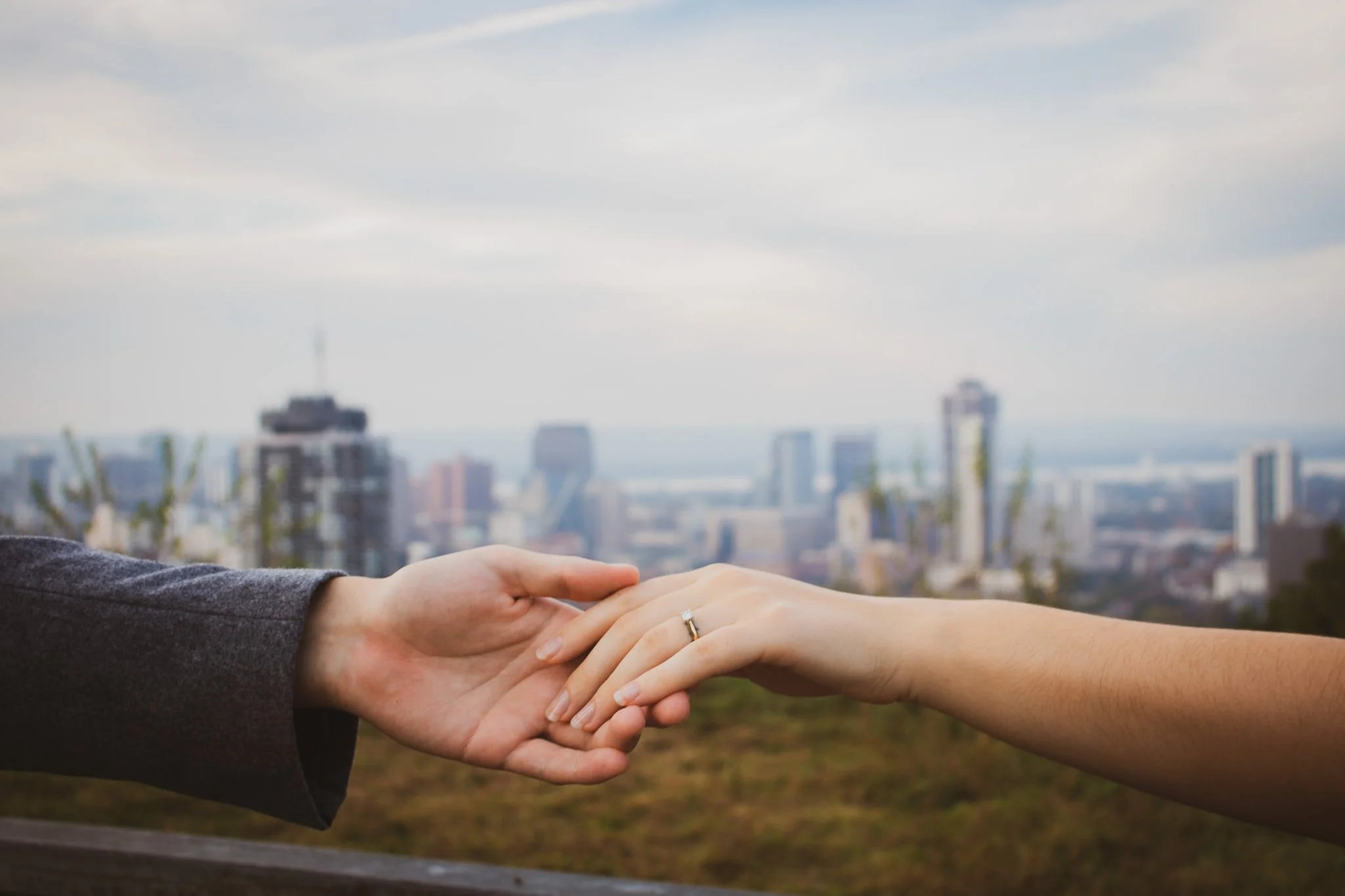 A couple holding hands with a city skyline in the background, the woman wearing a wedding ring.