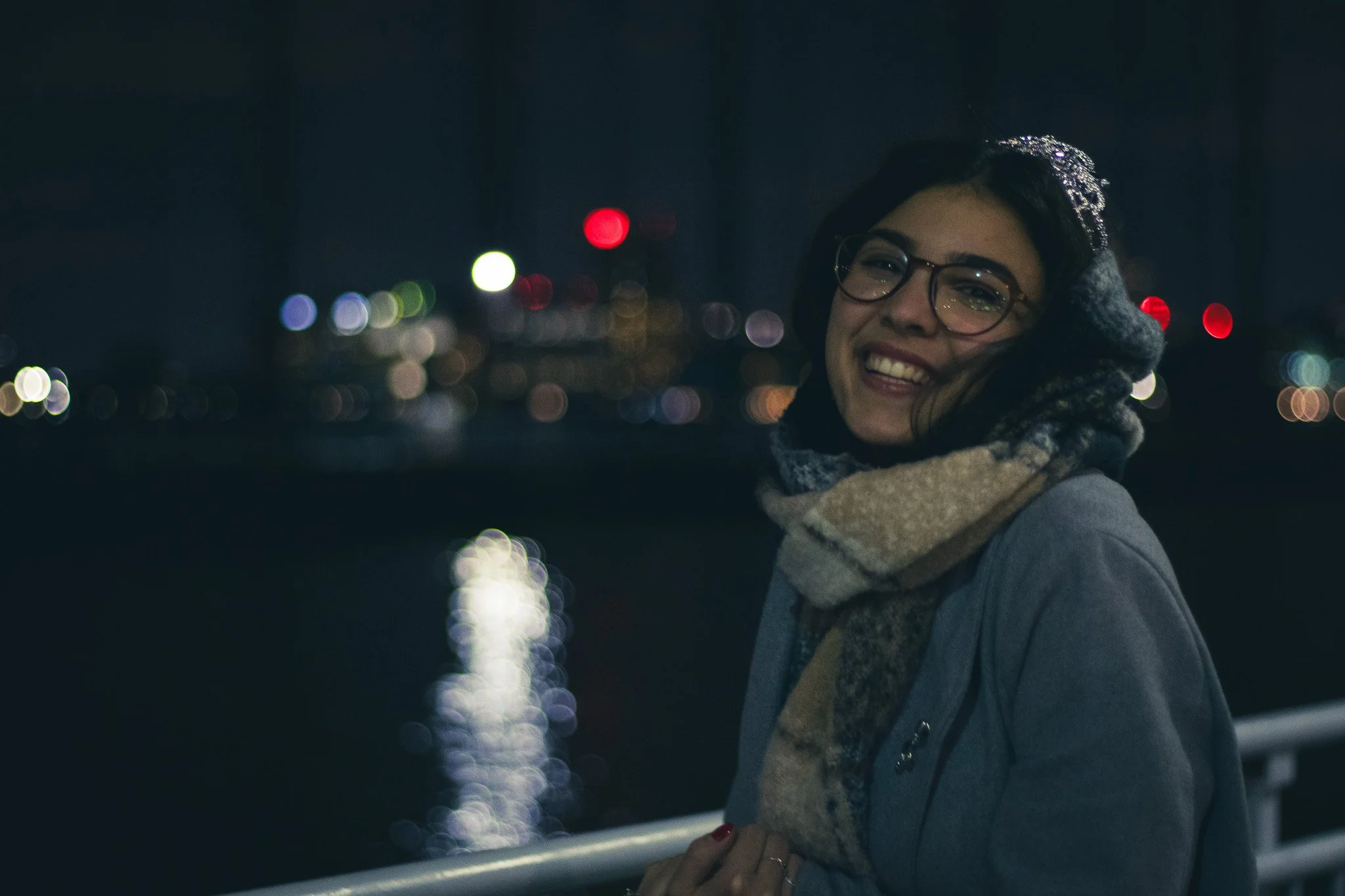 A smiling woman with glasses, wearing a dark coat, scarf, gloves, and a headband with rhinestones, outdoors at night with city lights reflecting on water in the background.