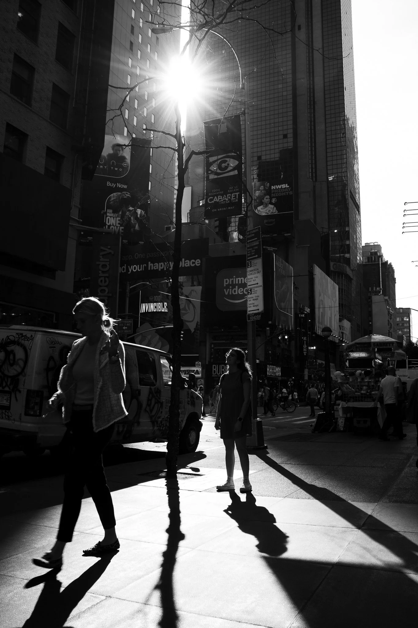 A busy city street scene with pedestrians walking, cars, and tall buildings. The sun shines brightly, creating strong shadows and glare. There are various billboards and storefront signs visible.