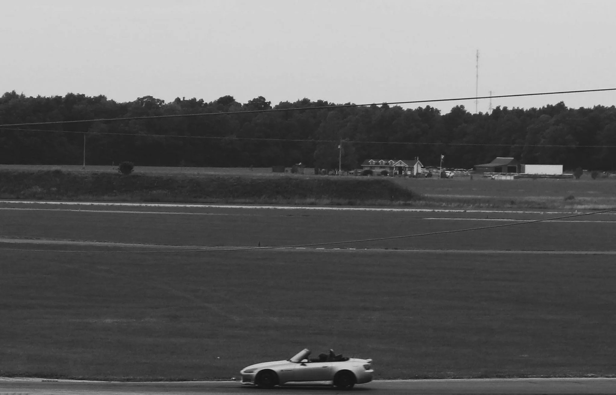 A black and white photo of a highway with a sports car driving past and a row of buildings and trees in the background.
