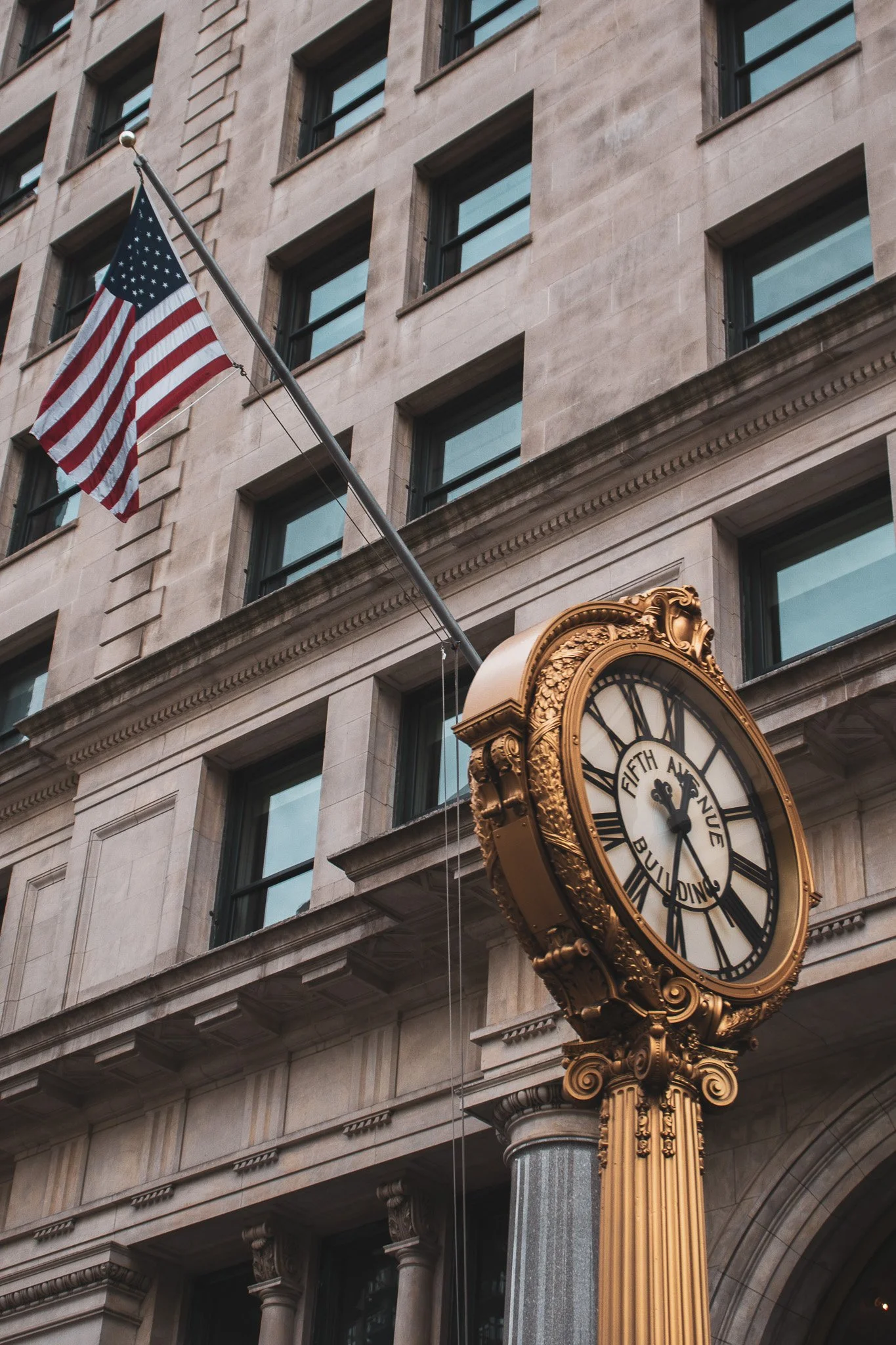An ornate clock with gold embellishments mounted on a decorative column outside a building with beige stone walls and multiple rectangular windows. An American flag is attached to a pole extending from the building.