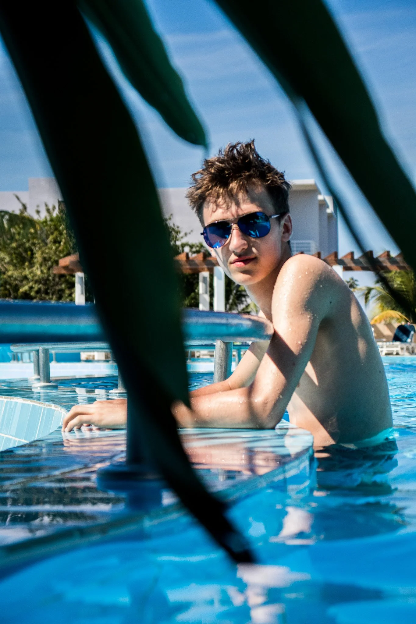 A young man with messy hair wearing reflective sunglasses sitting at the edge of a swimming pool, with palm trees, rooftops, and a bright blue sky in the background, partially framed by green leaves.