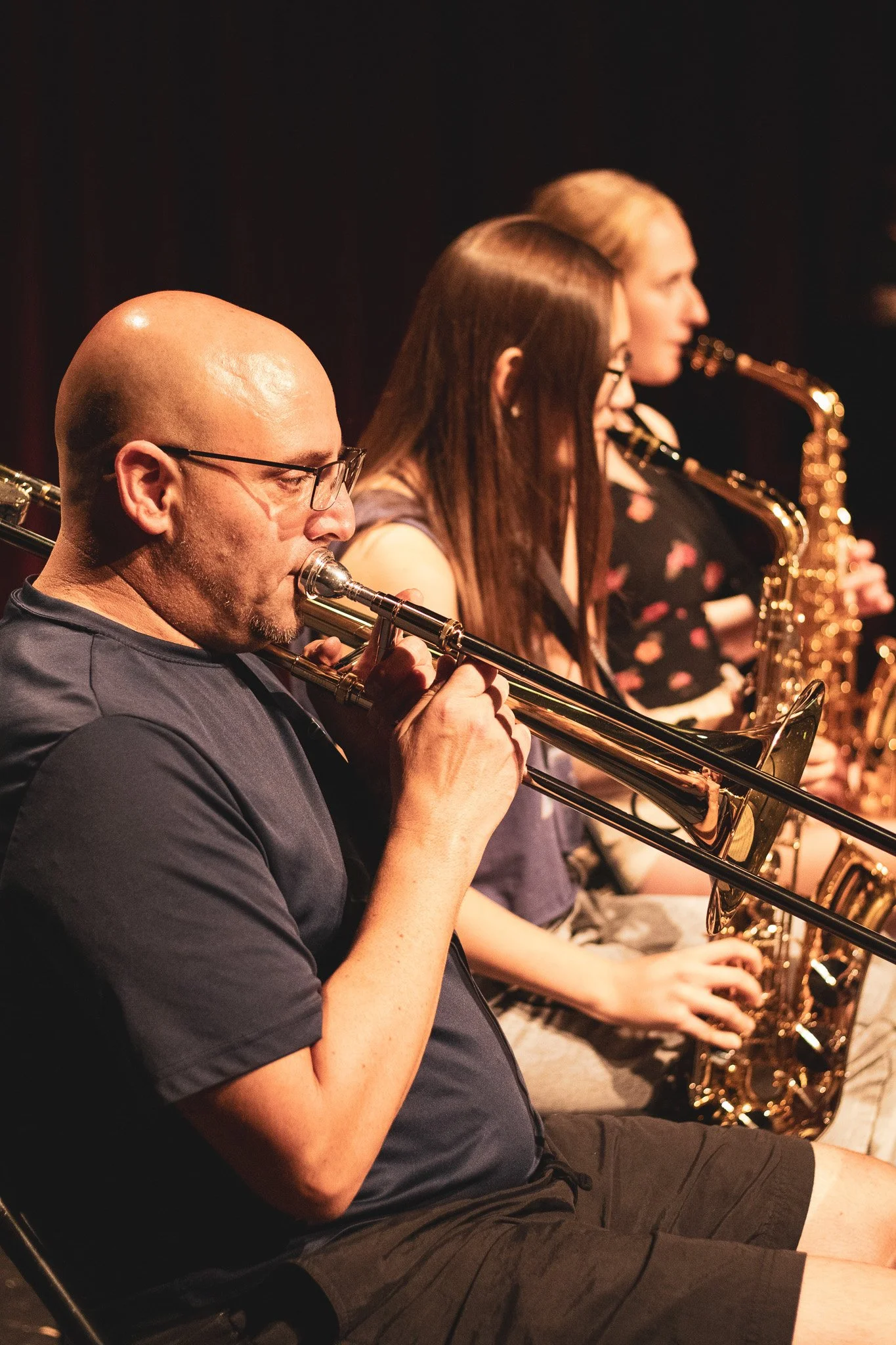 Three musicians playing wind instruments, seated in a row during a performance, with a dark background.