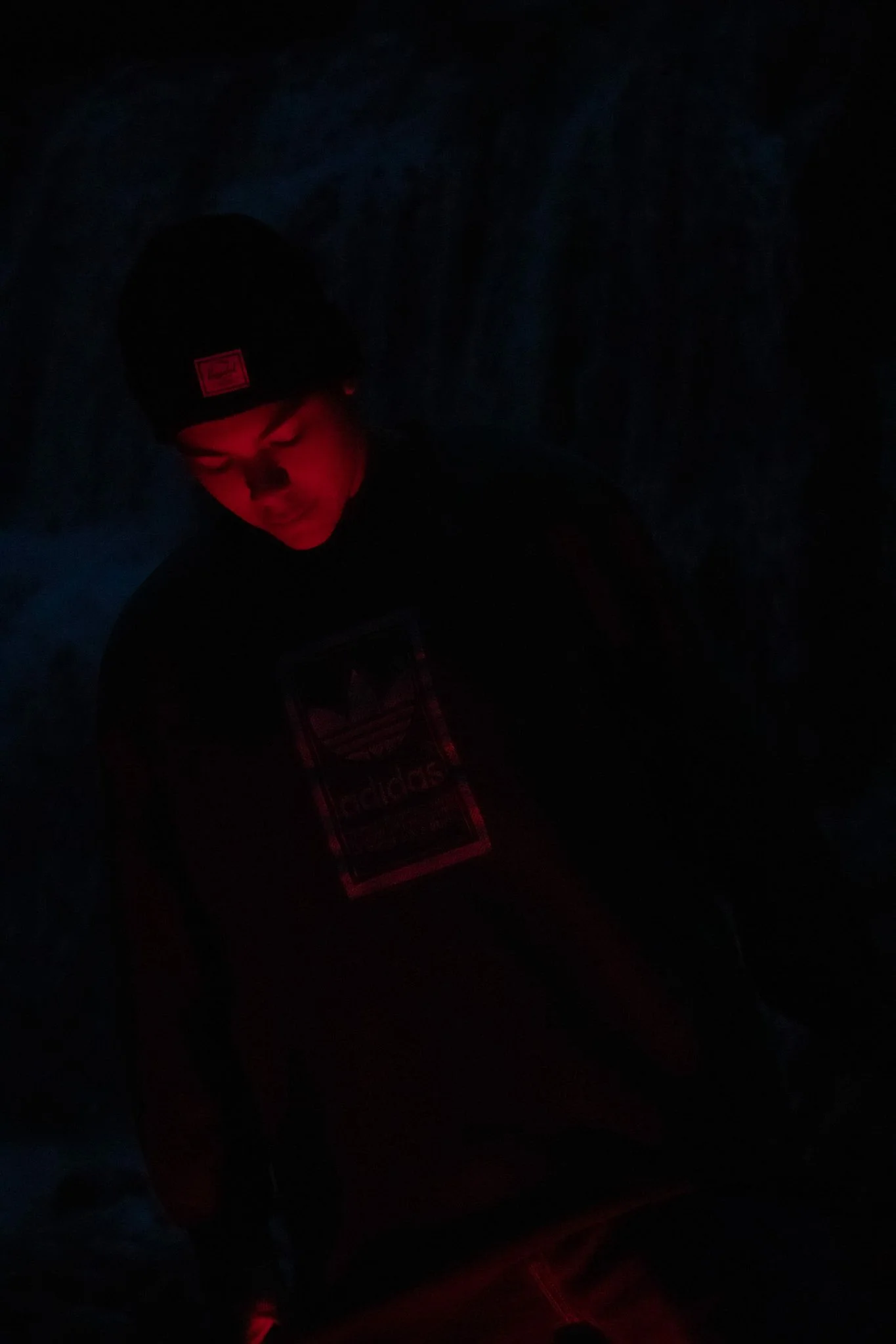 A person wearing a black beanie with a red logo and a dark hoodie, illuminated by red light, in a dark environment.