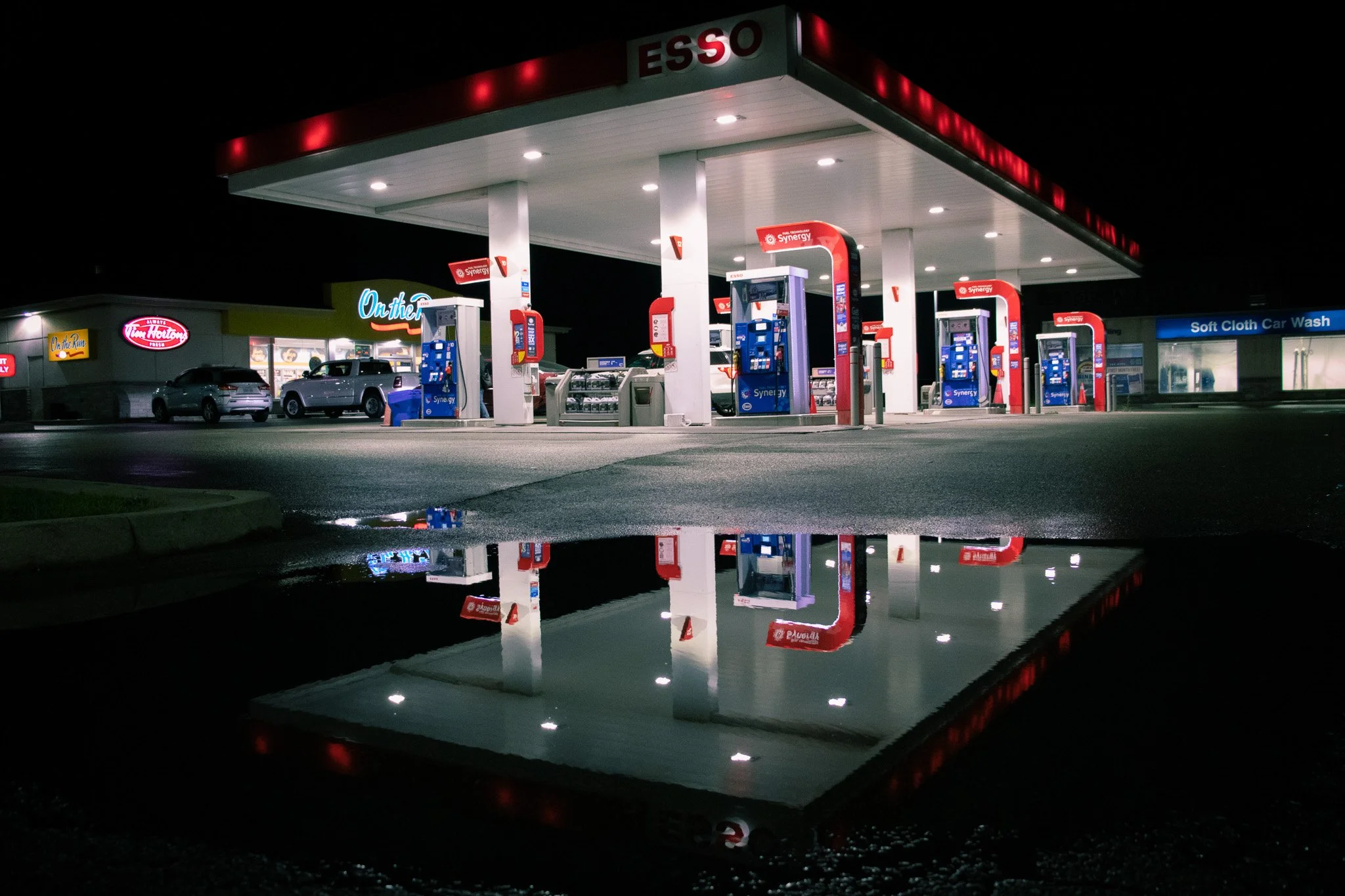 Nighttime view of an Esso gas station with illuminated pumps and signage, with parked cars and a soft cloth car wash in the background, and reflections on a wet surface.