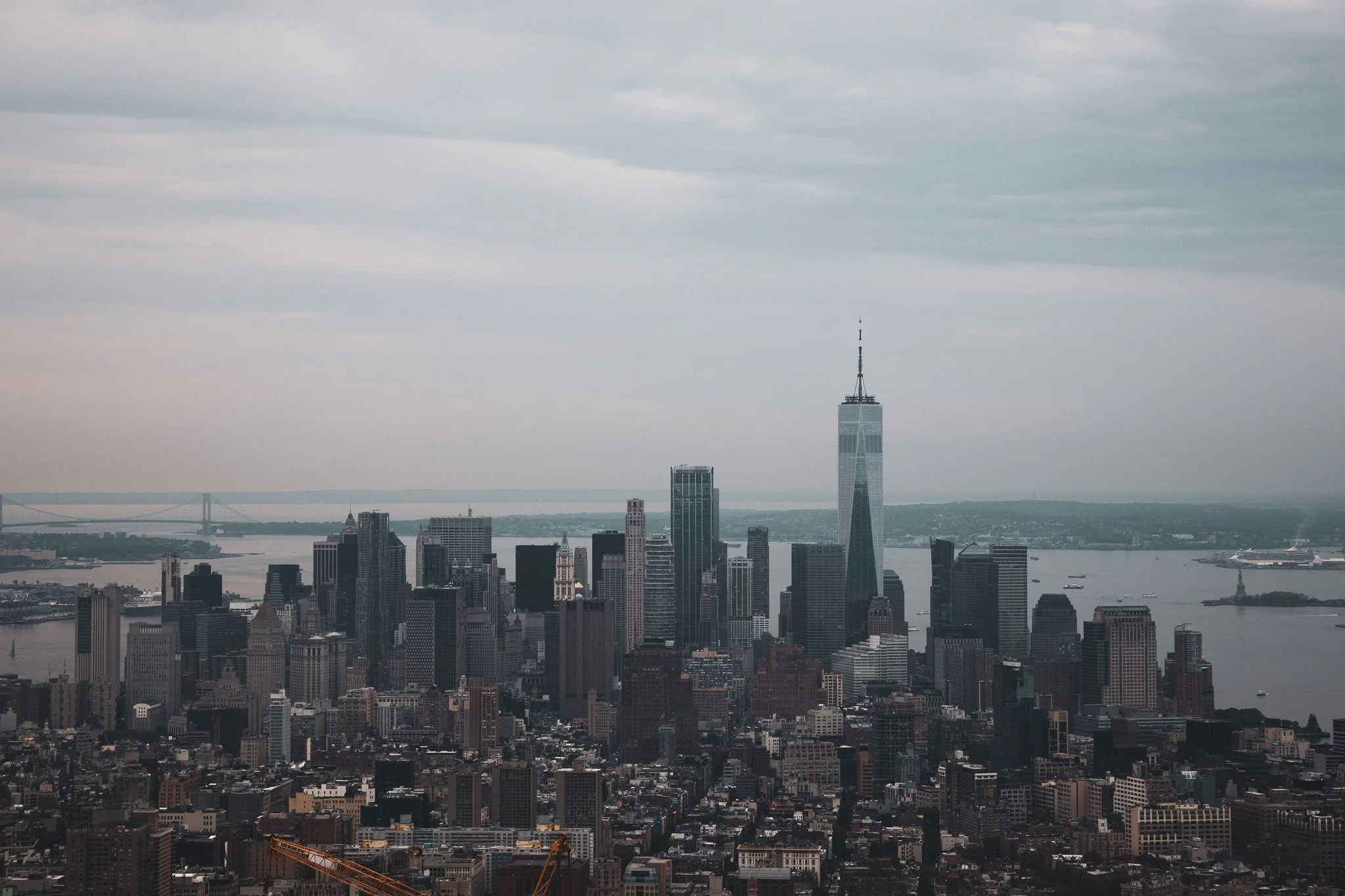 Overcast city skyline with tall skyscrapers, including One World Trade Center, and a river with boats in the background.
