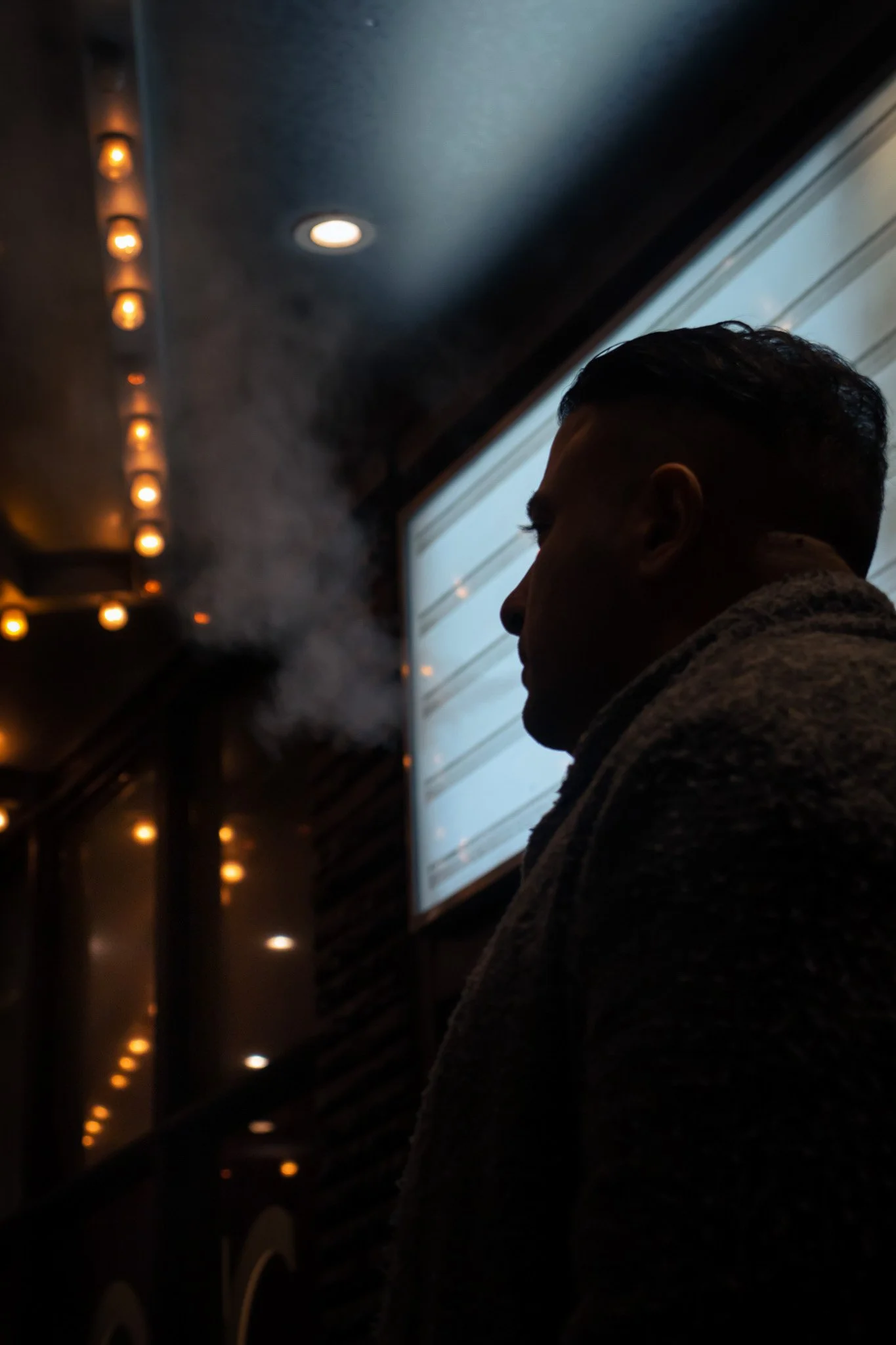 Silhouette of a person's profile next to a window with natural light, in a dimly lit room with warm ceiling lights.