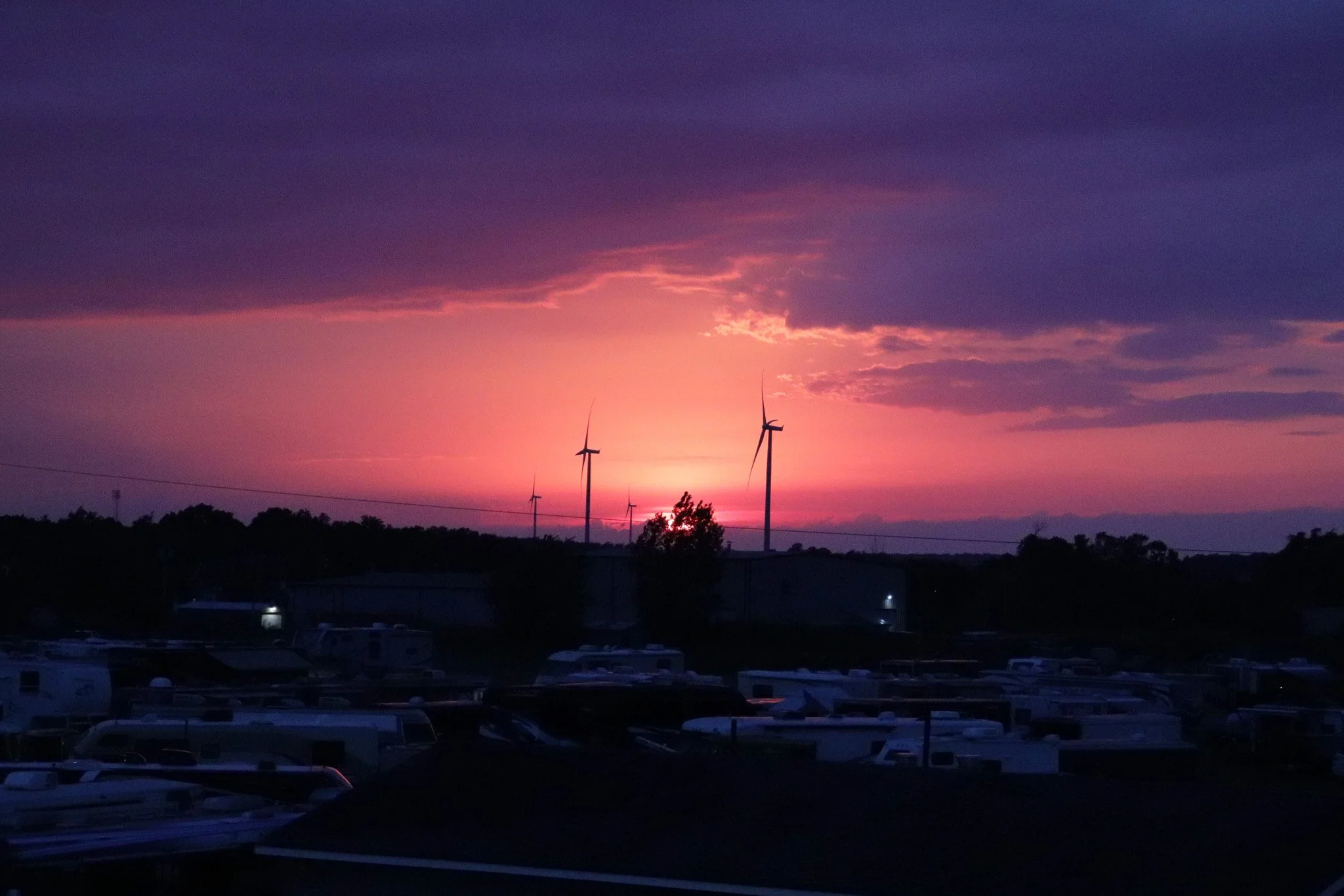 A sunset or sunrise sky with purple, pink, and orange hues, silhouetted wind turbines, trees, and parked cars below.