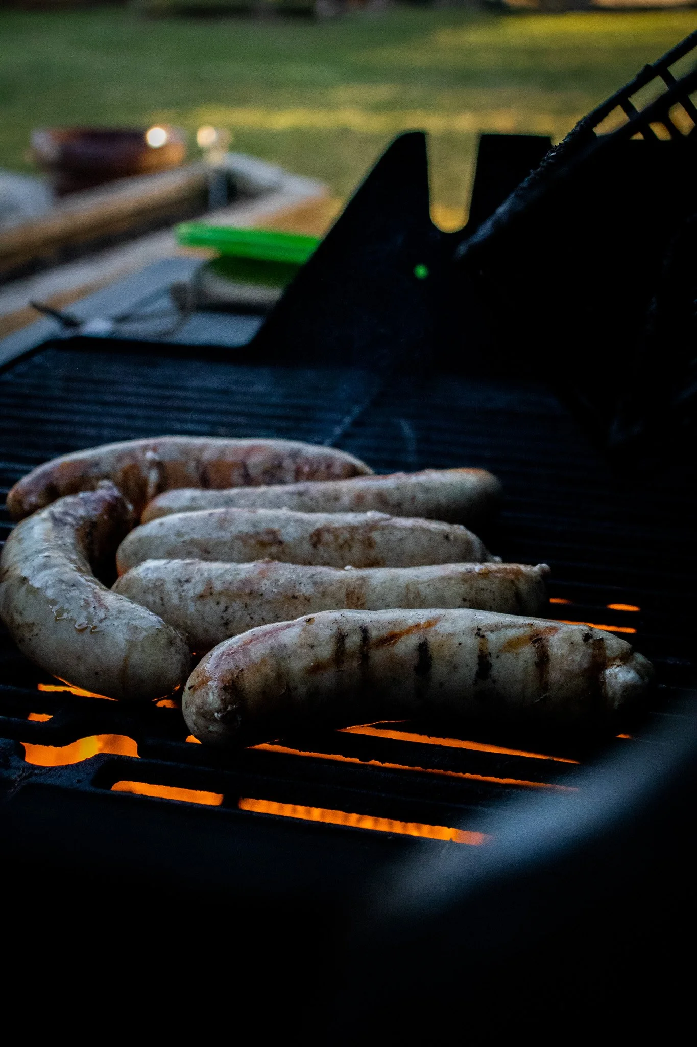 Raw sausages cooking on a grill over open flames with a backyard in the background.