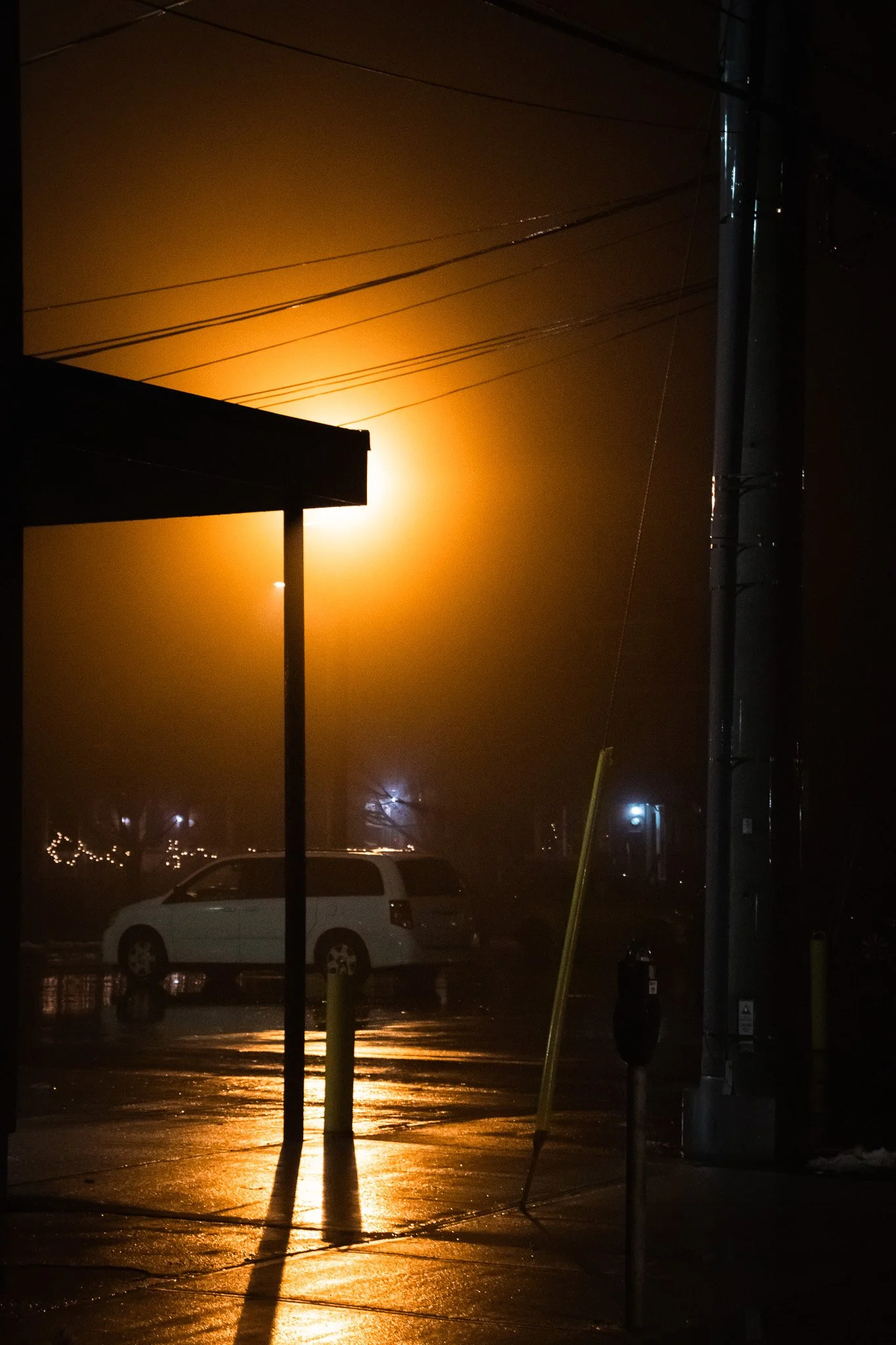 Night scene showing a parking lot with a white van, a traffic light, utility poles, power lines, and cars in the background. The sky is illuminated with bright orange light, possibly from a fire or intense street lighting, creating a reflective glow 