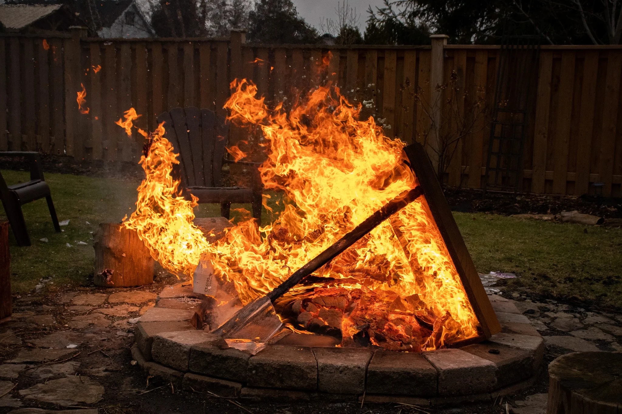 A backyard fire pit with flames burning, surrounded by patio stones, with a wooden Adirondack chair nearby, enclosed by a wooden fence during dusk or early evening.