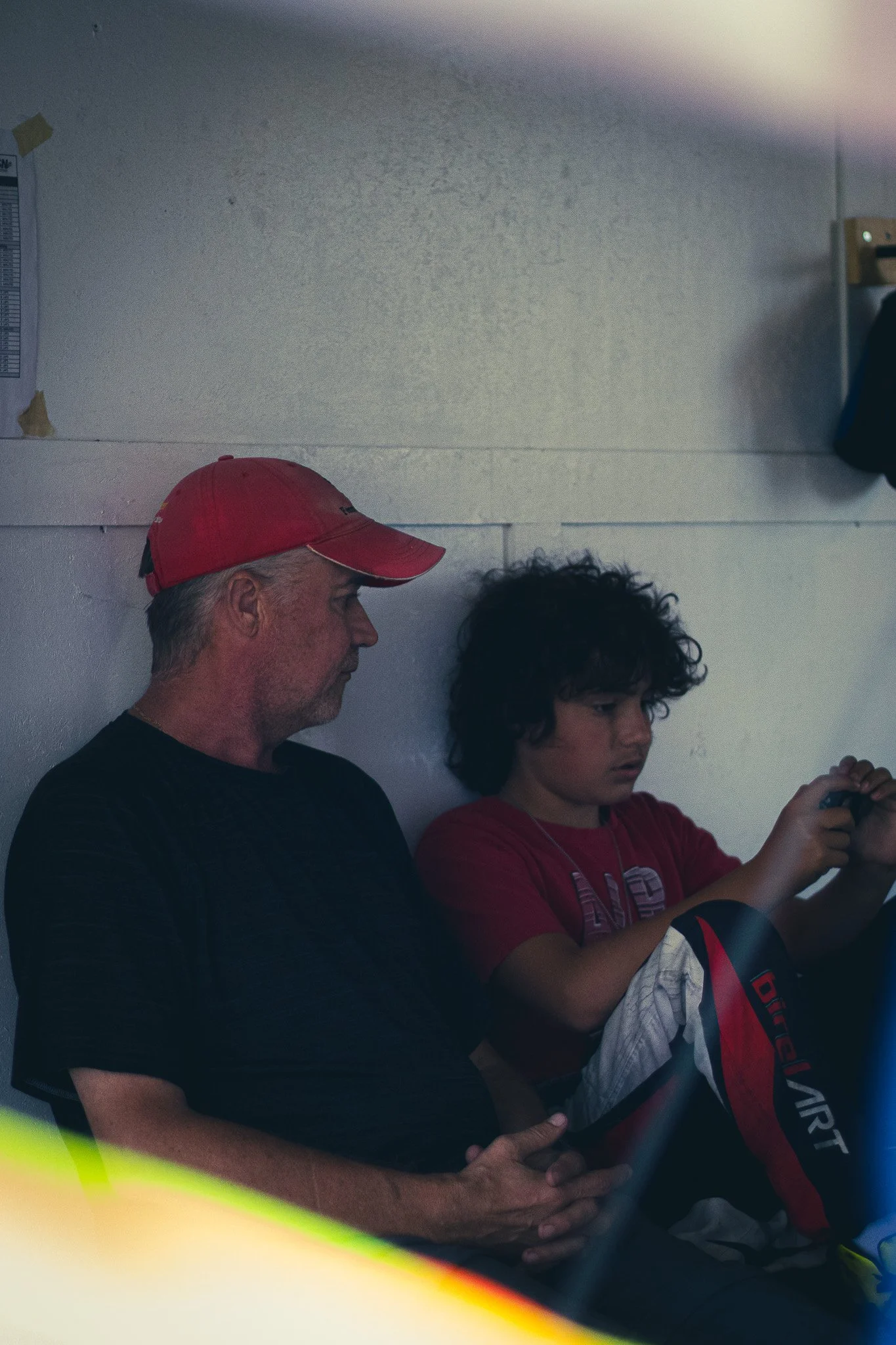 Two men sitting against a white wall, one older with a red cap and the other younger with curly hair, looking at a device.