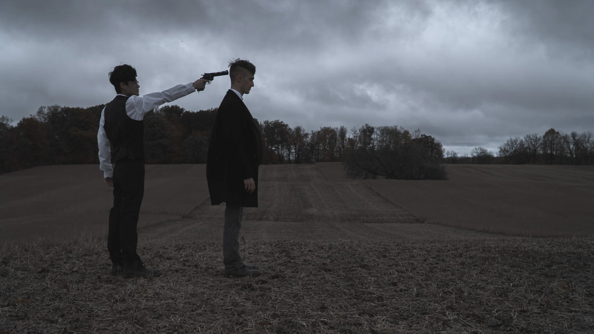 A person in a suit stands in a field with their back to the camera while another person in formal attire points a gun at their head. The sky is overcast.