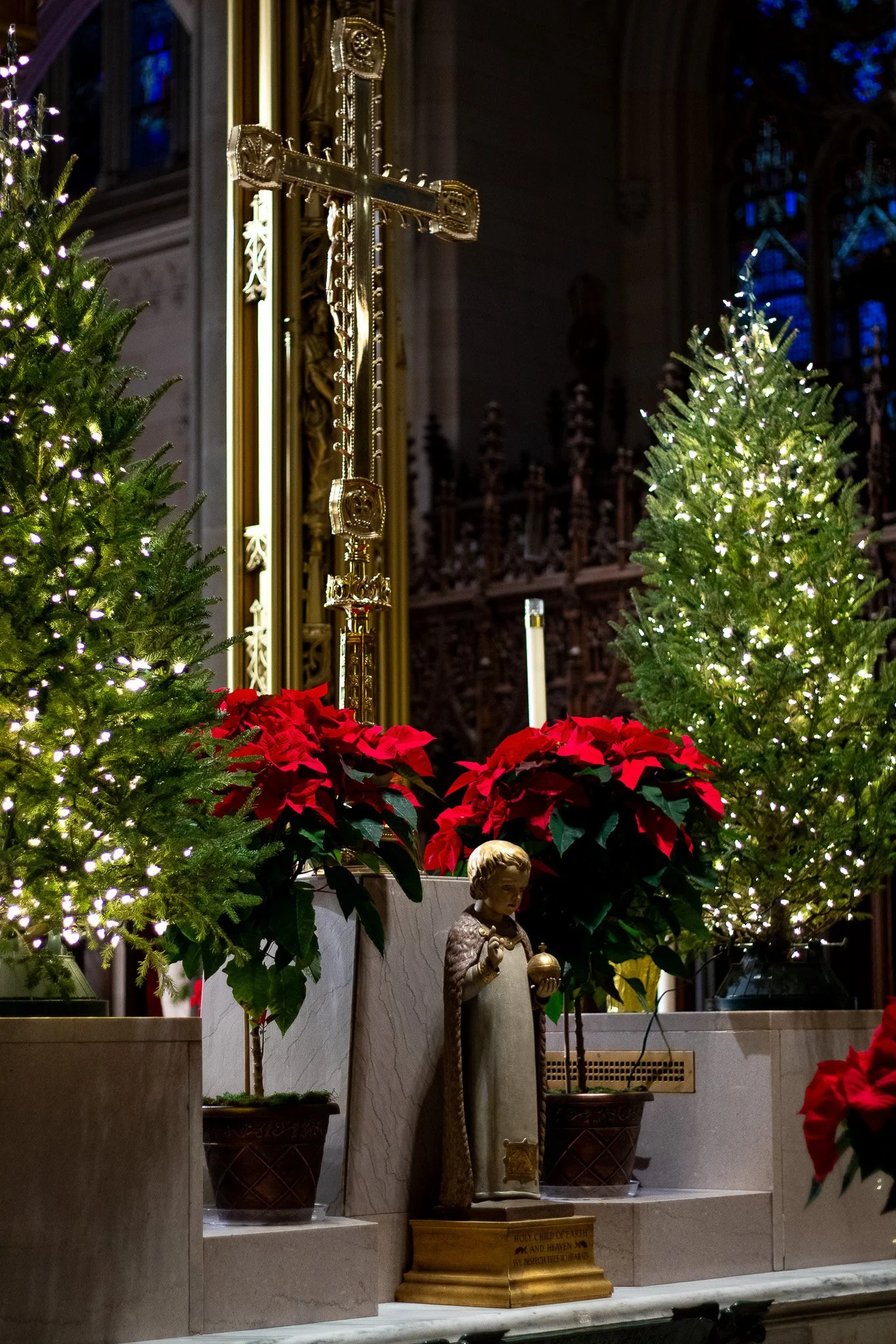 Christmas altar with two decorated Christmas trees, red poinsettias, a statue of a child holding an orb, and a gold crucifix, inside a church with gothic stained glass windows.