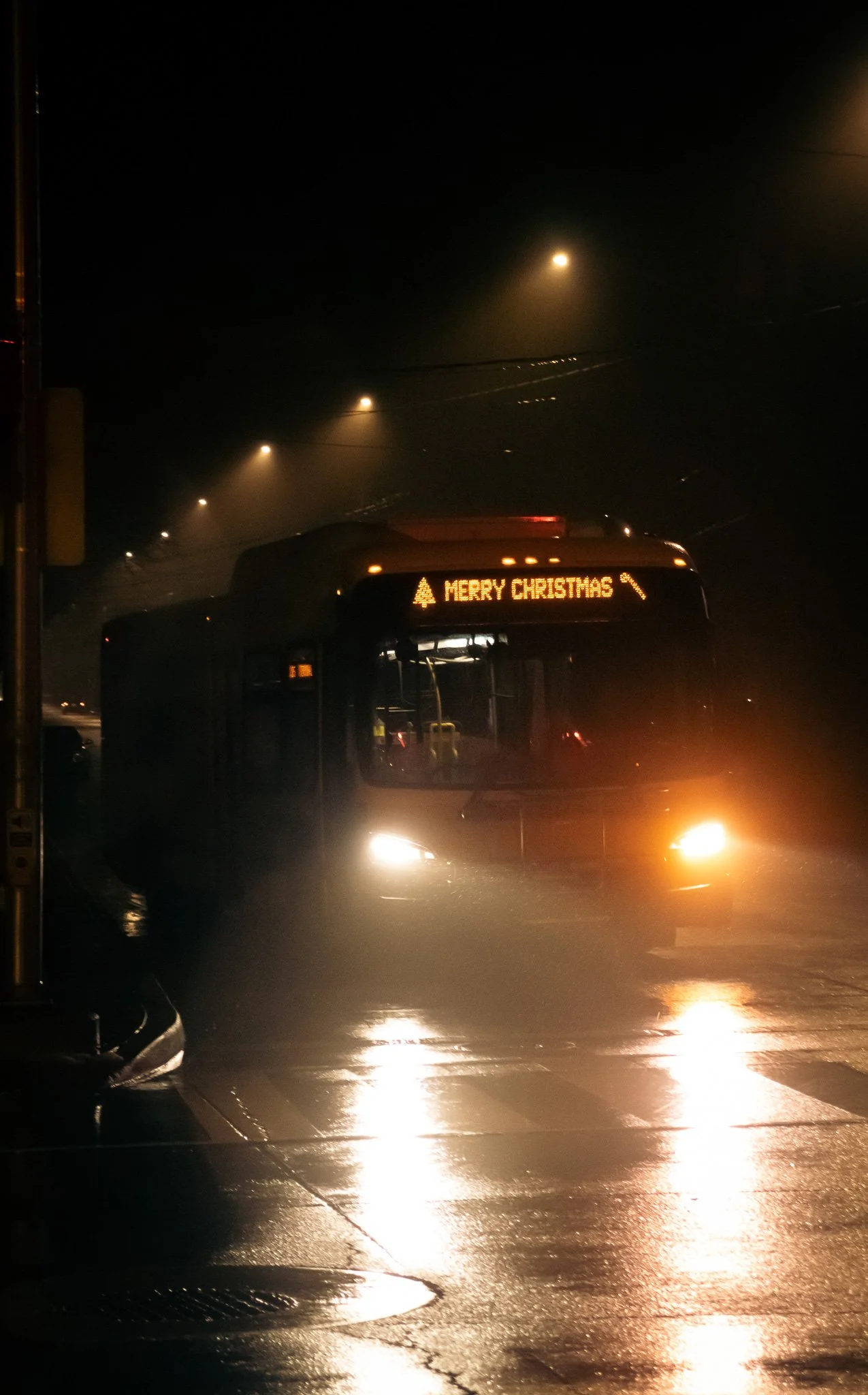 Nighttime scene on a rainy street with wet pavement, illuminated by streetlights. A bus displays a digital message saying 'Merry Christmas' with a Christmas tree icon.