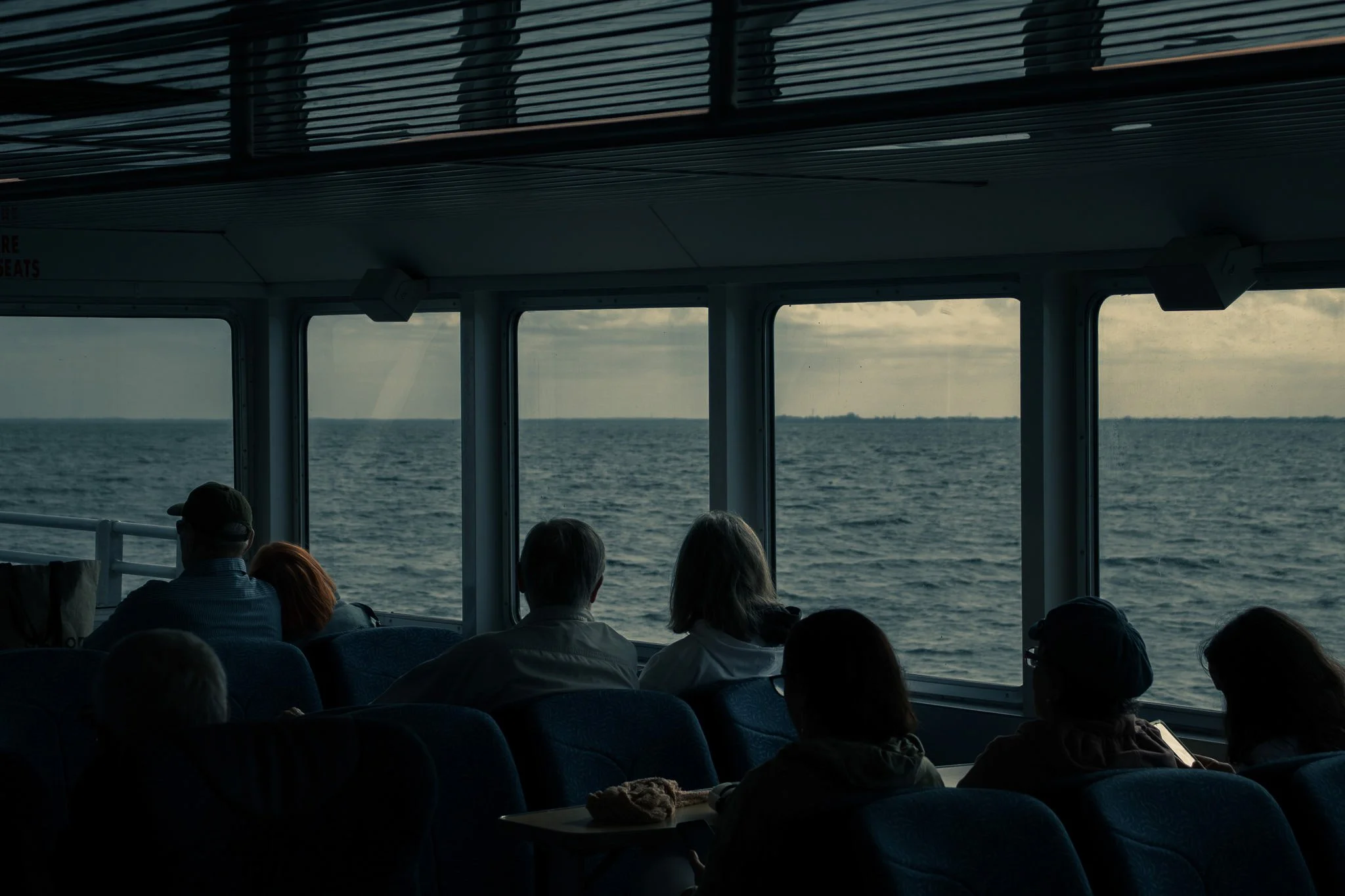 View of ocean through windows on a ferry or cruise ship, with passengers seated inside, some looking out and others possibly using devices, under a dark ceiling with light fixtures.