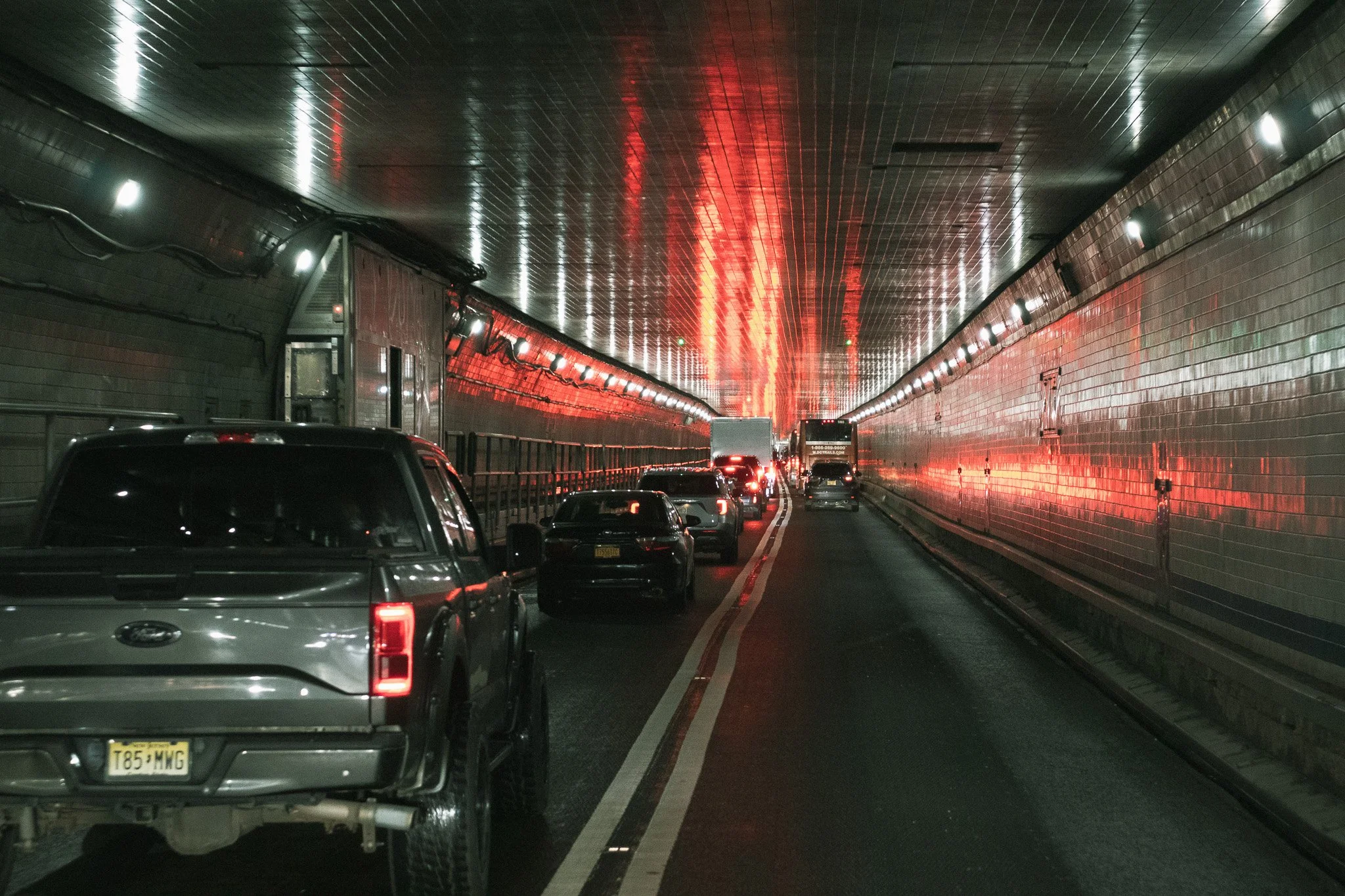 Vehicles driving through a tunnel with reflective tiled walls and ceiling, illuminated by bright white and red lights.