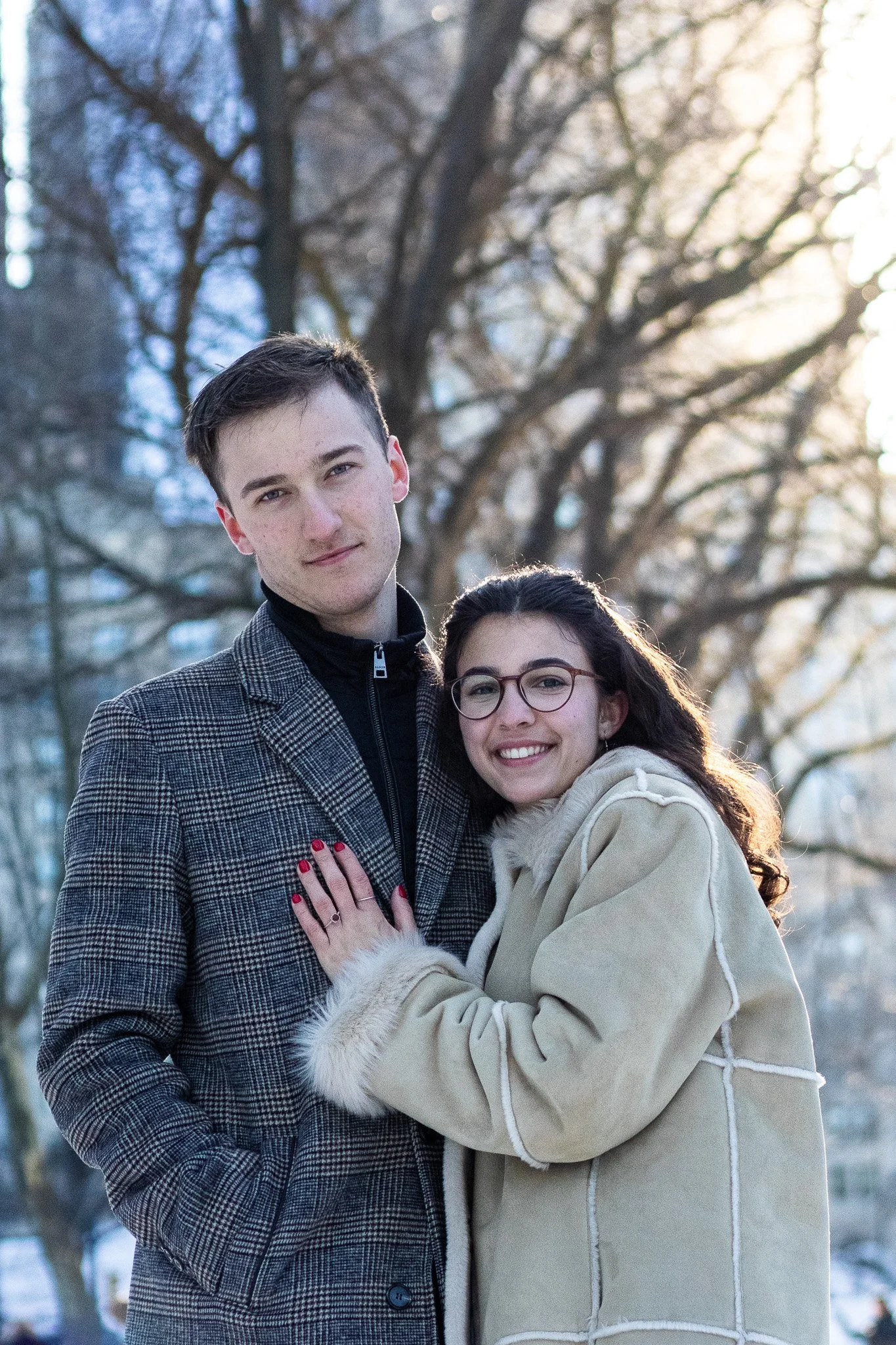A young man and woman standing outdoors in winter, close together, smiling, with leafless trees in the background. The man is wearing a plaid blazer and the woman is wearing glasses and a light-colored coat.
