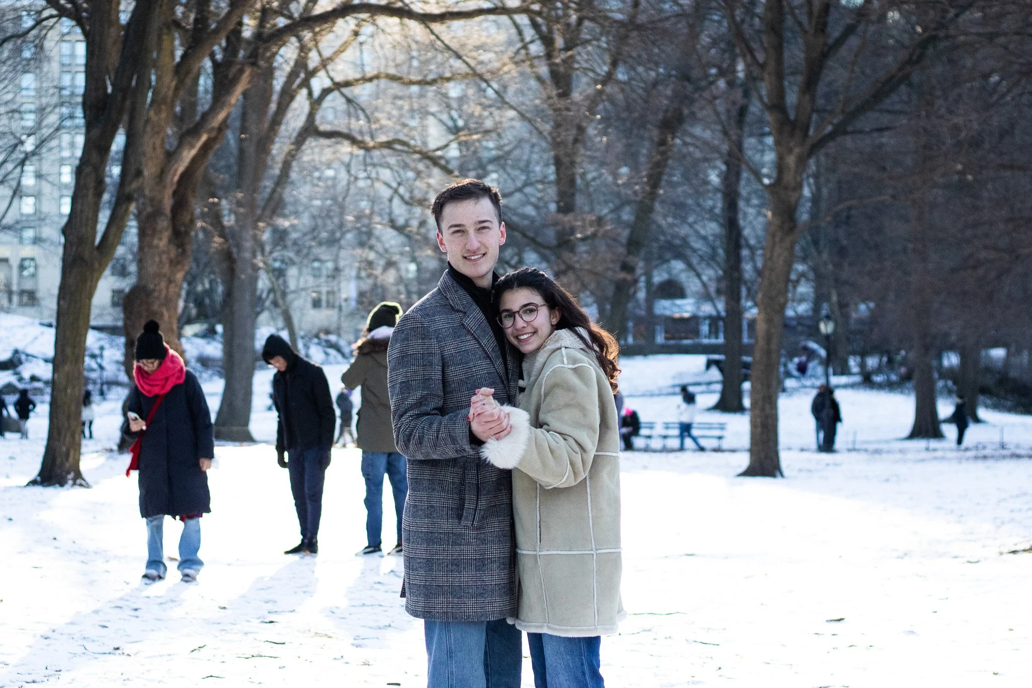 A young man and woman are smiling and dancing together in a snowy park, surrounded by leafless trees, with other people in the background.