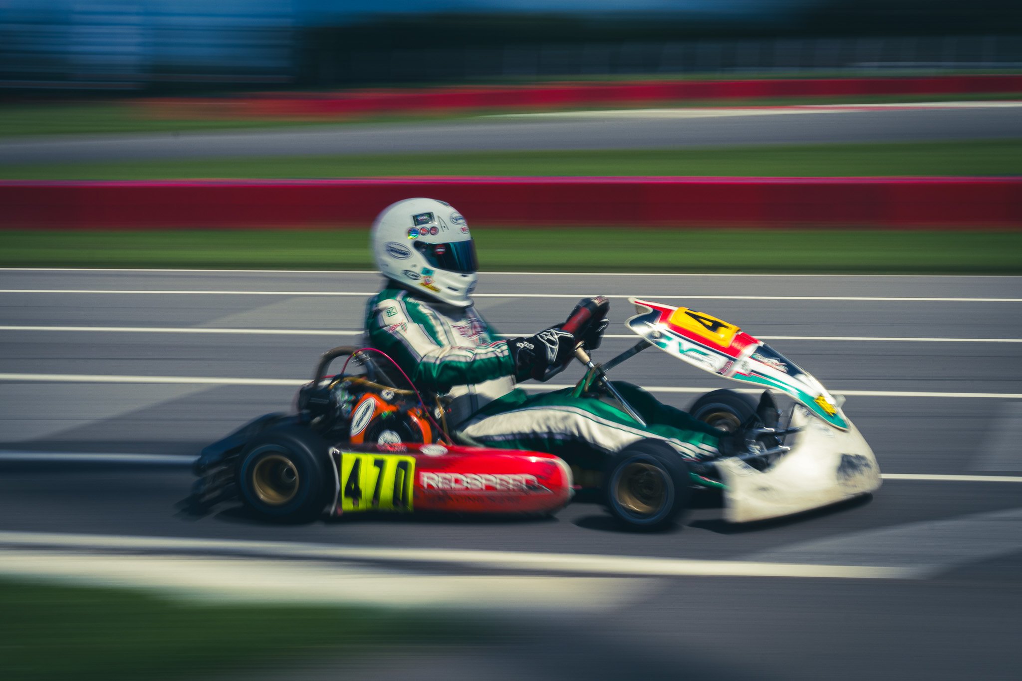Two go-kart racers competing on a track, with motion blur indicating high speed. One racer wears a white helmet and green racing suit, while the other wears a red and black kart with the number 470.