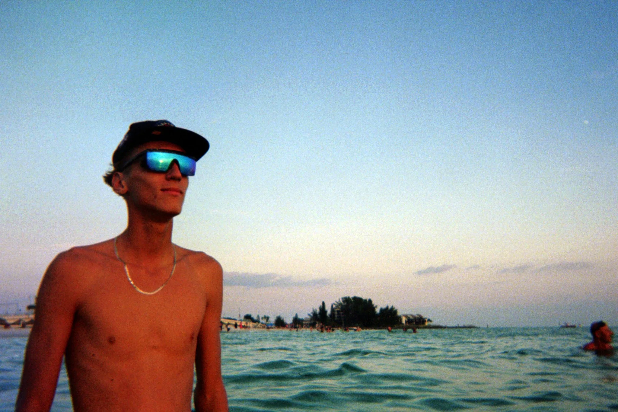A shirtless young man wearing sunglasses and a baseball cap standing near the water at the beach during sunset, with another person in the water and beach structures in the background.