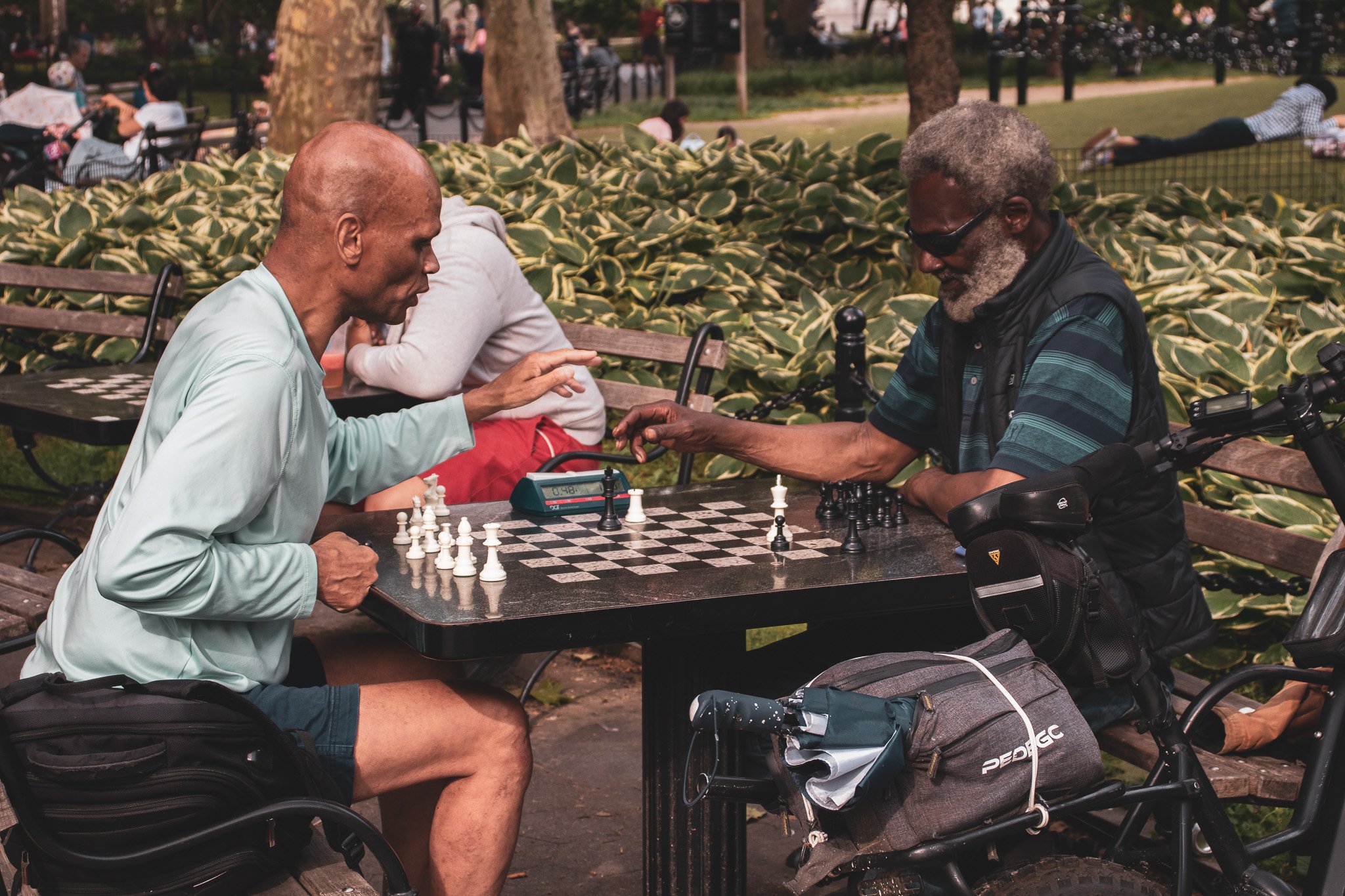 Two men playing chess outdoors in a park. One man is wearing a light-colored shirt and shorts, and the other is wearing a striped shirt, a vest, and sunglasses. There are bicycles and bags nearby, and other people are sitting and walking in the backg