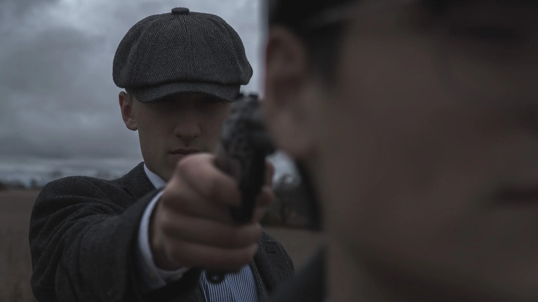 A man dressed in vintage clothing, wearing a flat cap and pointing a gun directly at the camera on a cloudy day by the coast.