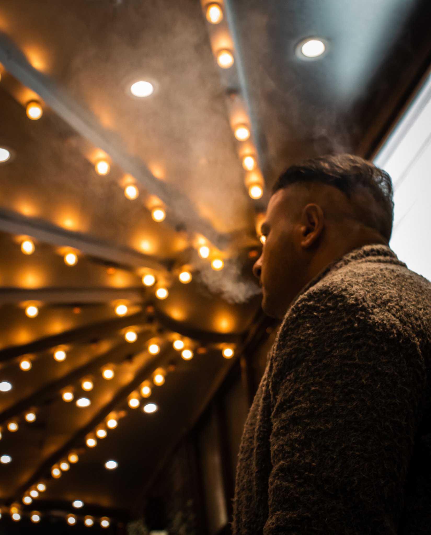 Person with short dark hair and dark skin, wearing a textured coat, under a ceiling with warm, yellowish string lights in an indoor setting.