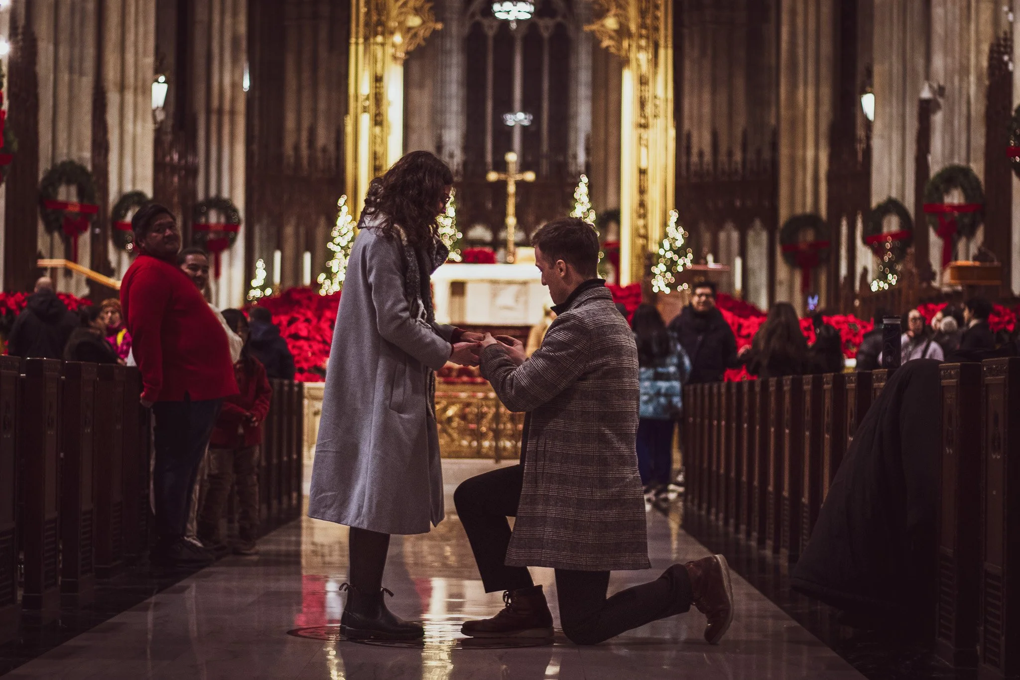 A woman proposing marriage to a man in a church decorated for Christmas. The man is kneeling, holding the woman's hands as she looks down at him, in front of an altar with a cross and Christmas trees.