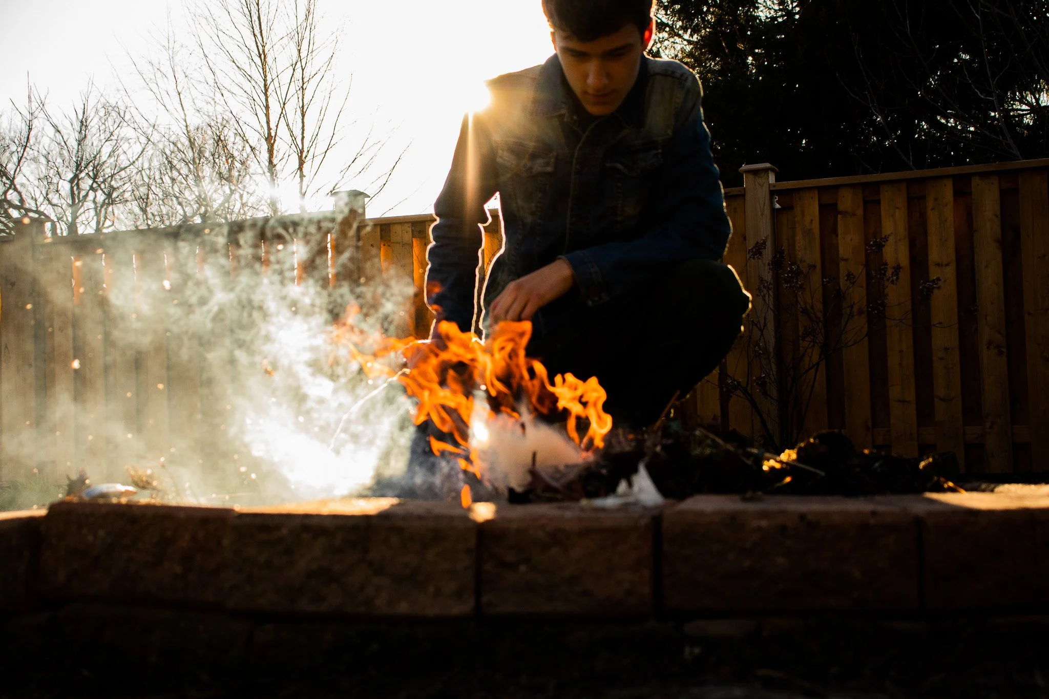 A young man crouched near a fire in a backyard during sunset, with leafless trees and a wooden fence in the background.