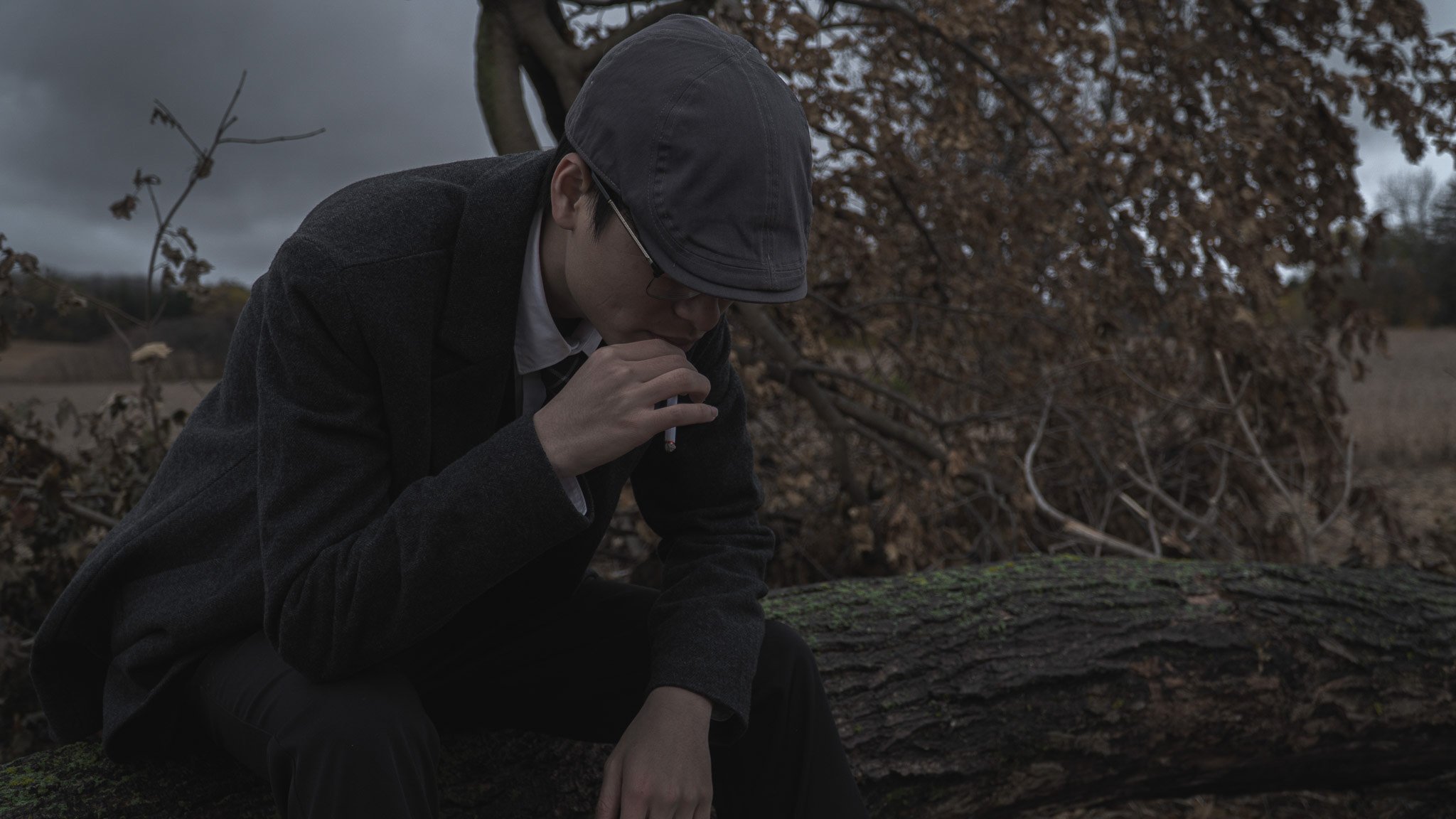 A person wearing a dark cap and glasses, dressed in a gray jacket, sitting on a log outdoors, smoking a cigarette. The background features a tree with brown leaves and cloudy sky, suggesting a cool or overcast day.