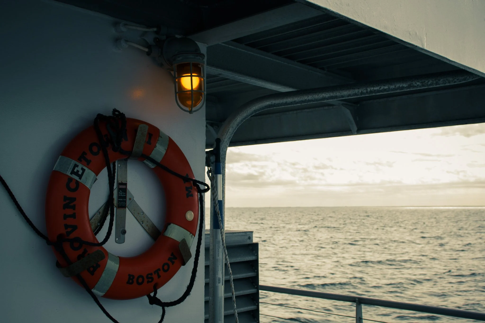 An orange life buoy with the words 'BOSTON' and 'NO' on it, mounted on a boat's white wall near a water body, with a yellow light fixture above.