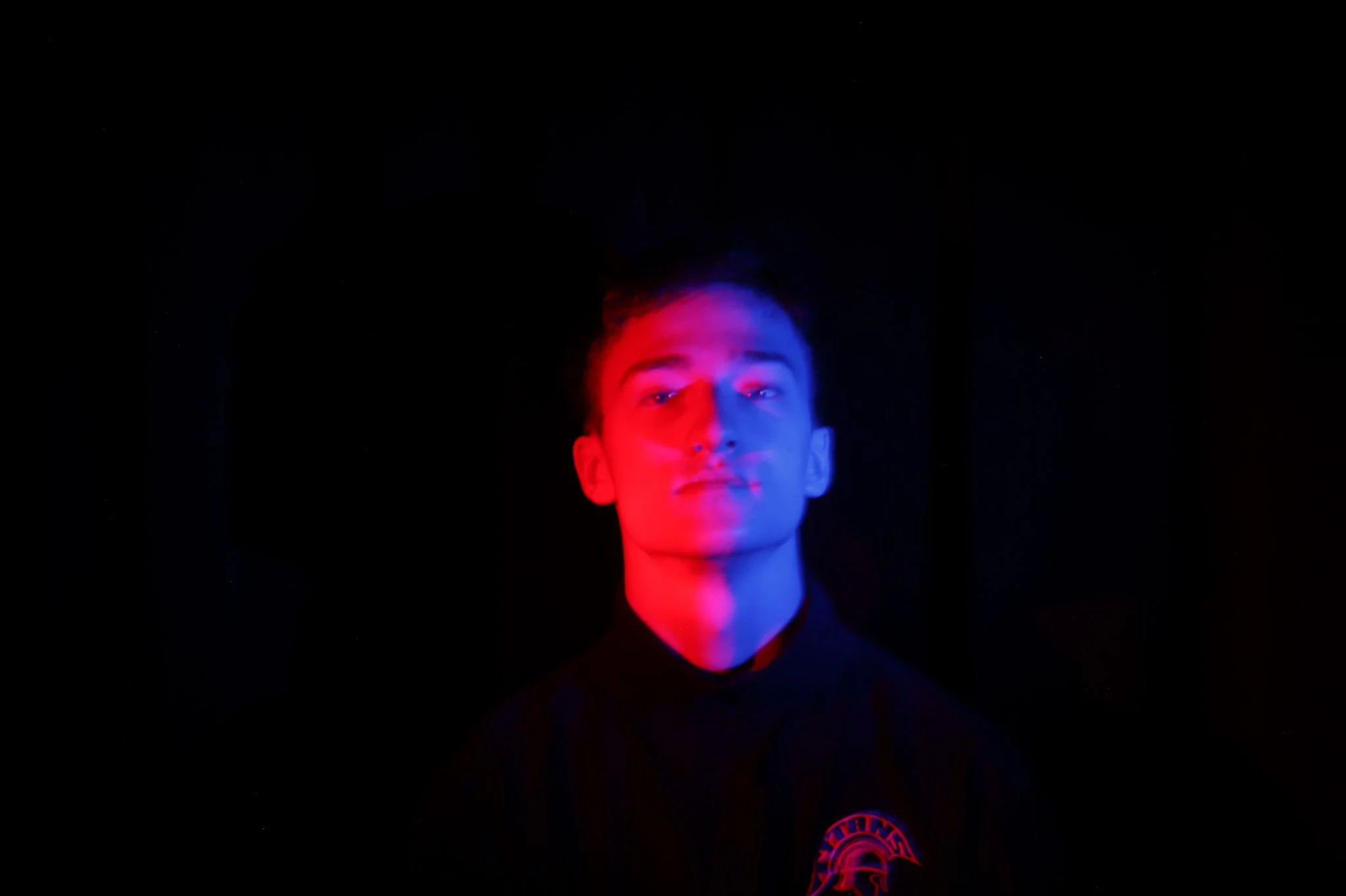 A person with short hair is illuminated with red and blue lighting, against a dark background.