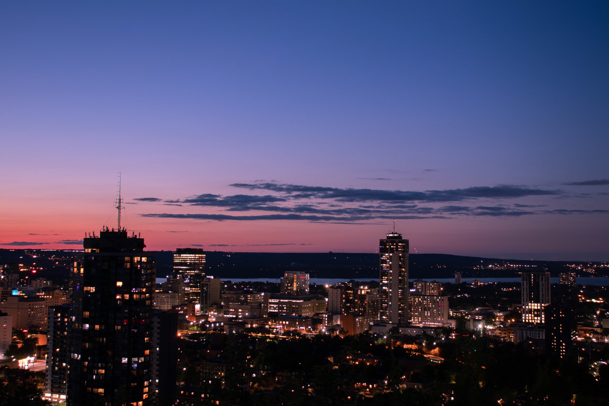 City skyline at dusk with high-rise buildings, a river in the background, and a sky painted with purple and orange hues.