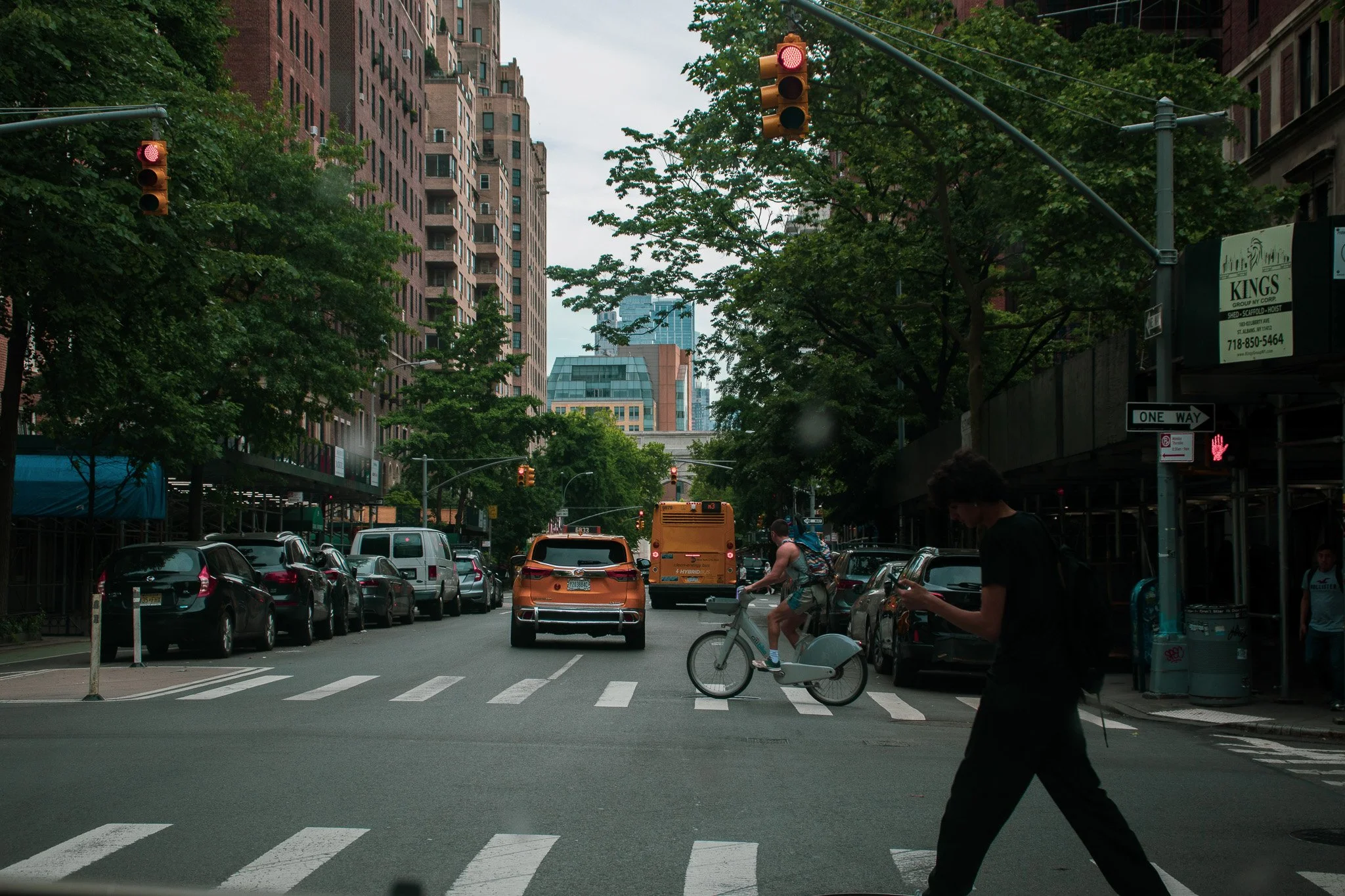 City street scene with red traffic lights, cars stopped at a crosswalk, people crossing the street, tall buildings, trees, and cloudy sky.