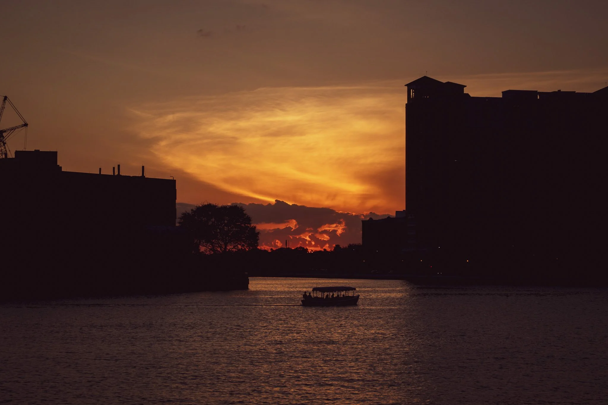 Silhouette of buildings and a crane against a vibrant orange and yellow sunset sky, with a river and a small boat in the foreground.