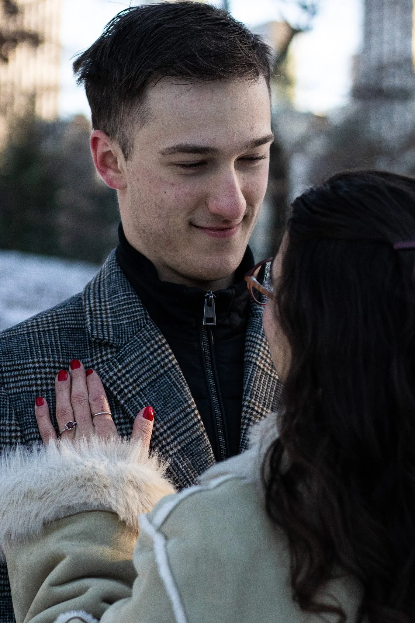 A young man with dark hair and fair skin smiling gently as he looks downward at a woman wearing glasses, who is touching his chest with her hand. The background is a blurry outdoor scene with some buildings and trees.