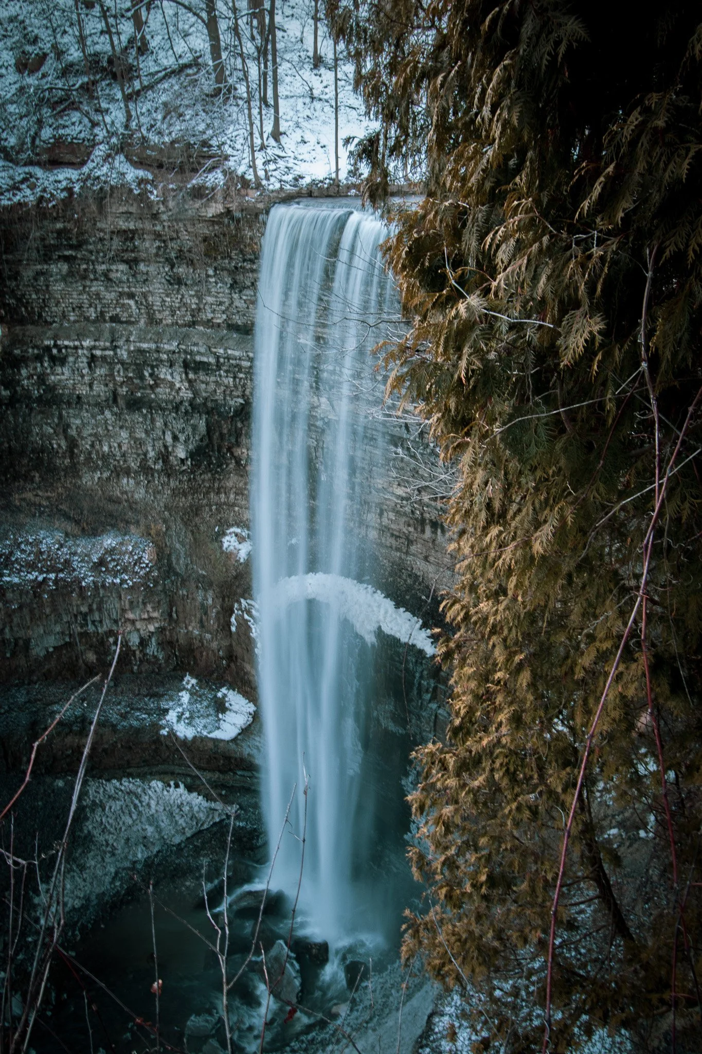 A waterfall falling into a river below, with snow on the ground and trees.