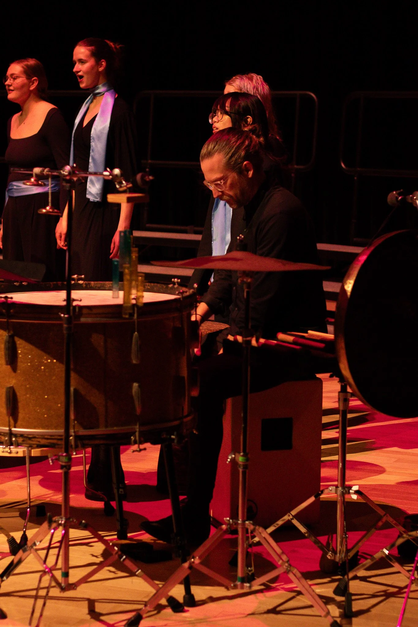 Musicians performing on stage with percussion instruments, people singing, and musical equipment in a concert hall.