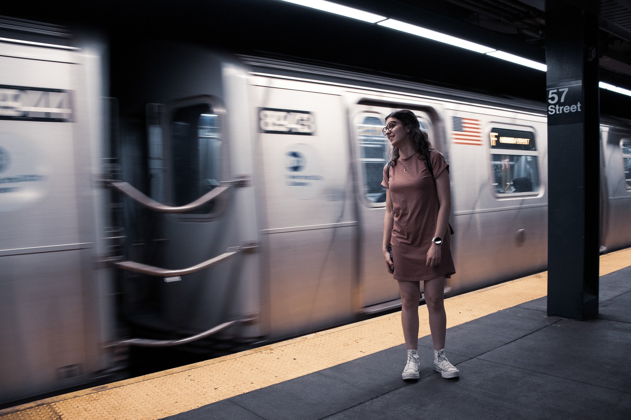 A woman with glasses, wearing a pink T-shirt dress and sneakers, stands on a subway platform next to a train in motion. The platform is marked with a yellow safety line. The station sign reads '57 Street.'
