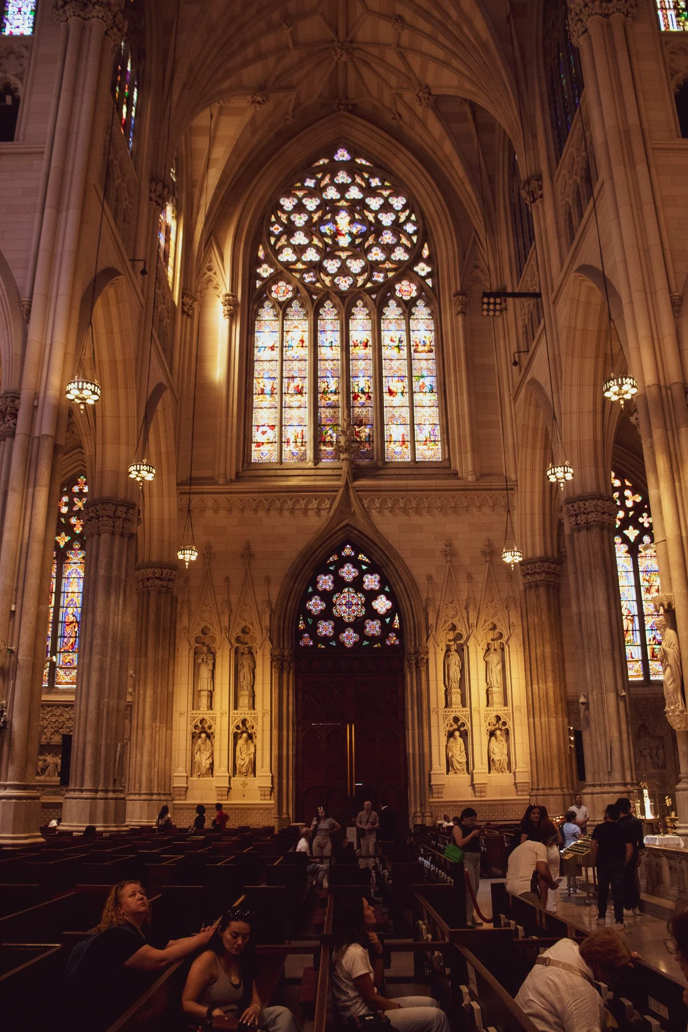Inside a Gothic-style cathedral with intricate stained glass windows, high vaulted ceilings, and stone sculptures. People are seated and standing, some near the altar area.