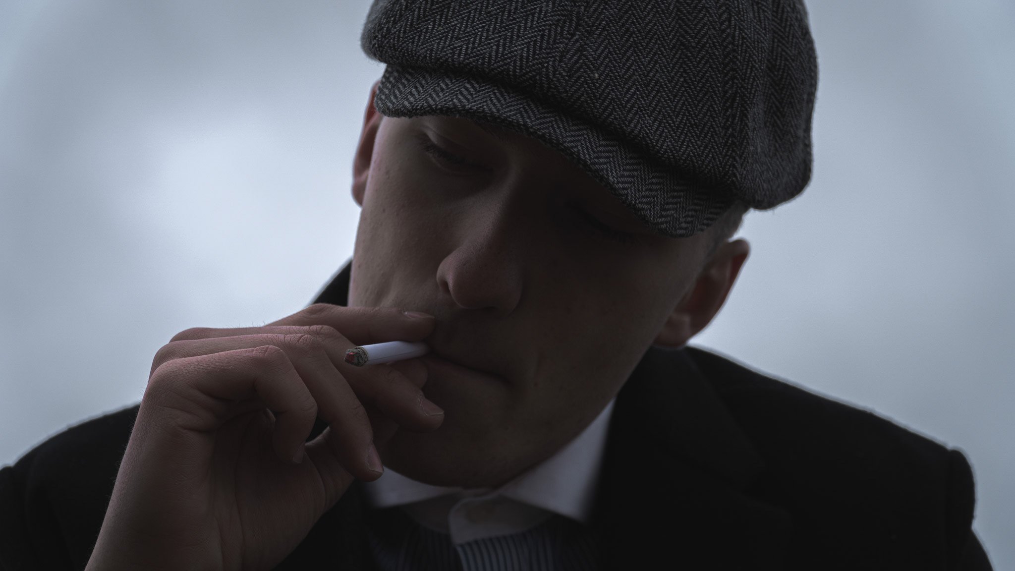 A young person wearing a dark blazer and a herringbone flat cap, smoking a cigarette against a cloudy sky.