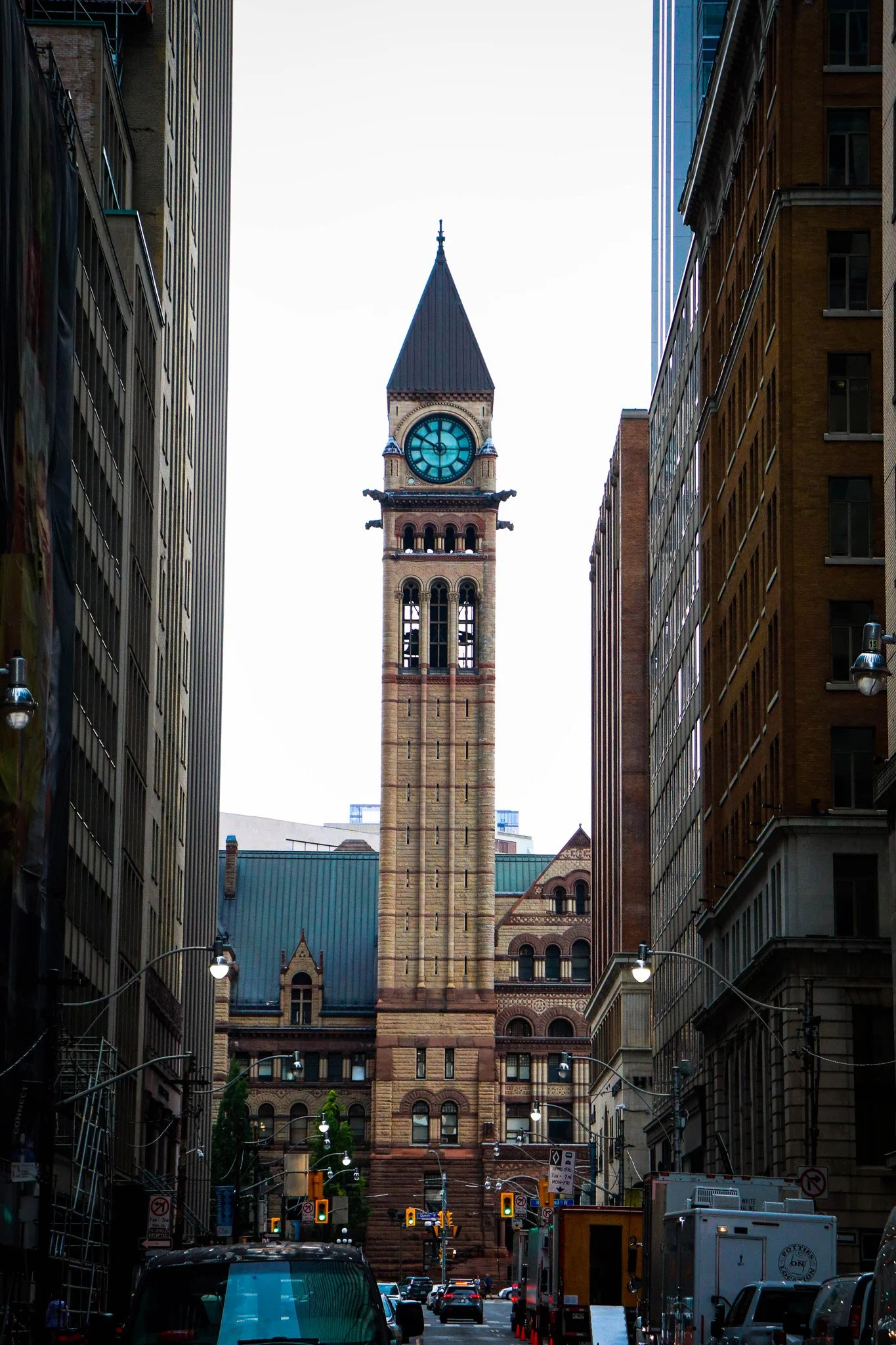 Tall clock tower with a pointed roof, located between two tall buildings in an urban area with traffic and streetlights.