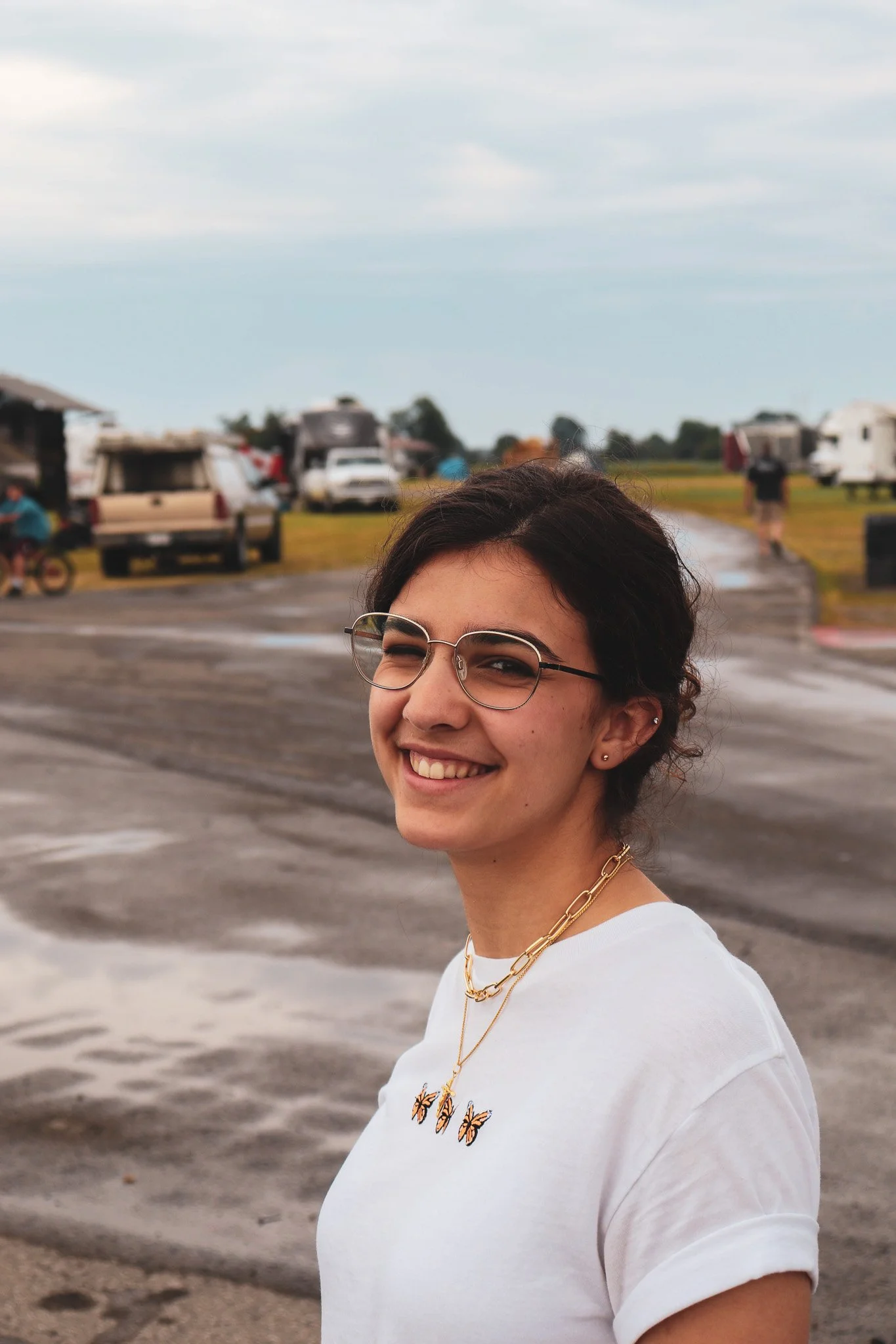 A woman wearing glasses and gold jewelry smiling outdoors with RVs and vehicles in the background on a cloudy day.