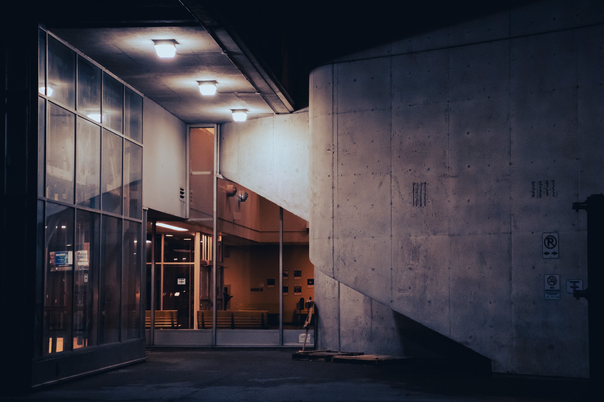 Nighttime scene of a modern building entrance with glass walls and a concrete wall, illuminated by ceiling lights. The setting appears to be an urban or institutional area with a sidewalk and signage.
