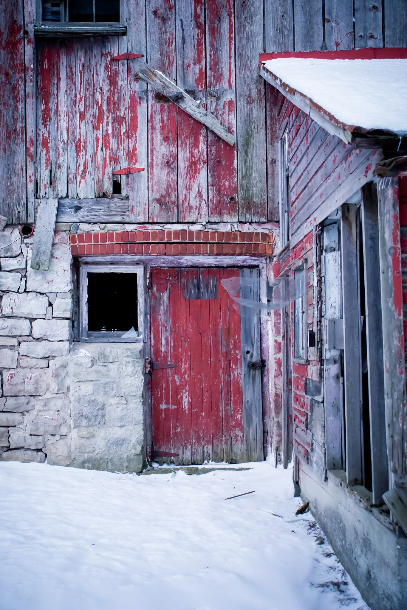 Old, weathered red wooden barn with a snow-covered roof, a small window, and a closed door, with snow on the ground and a stone foundation.