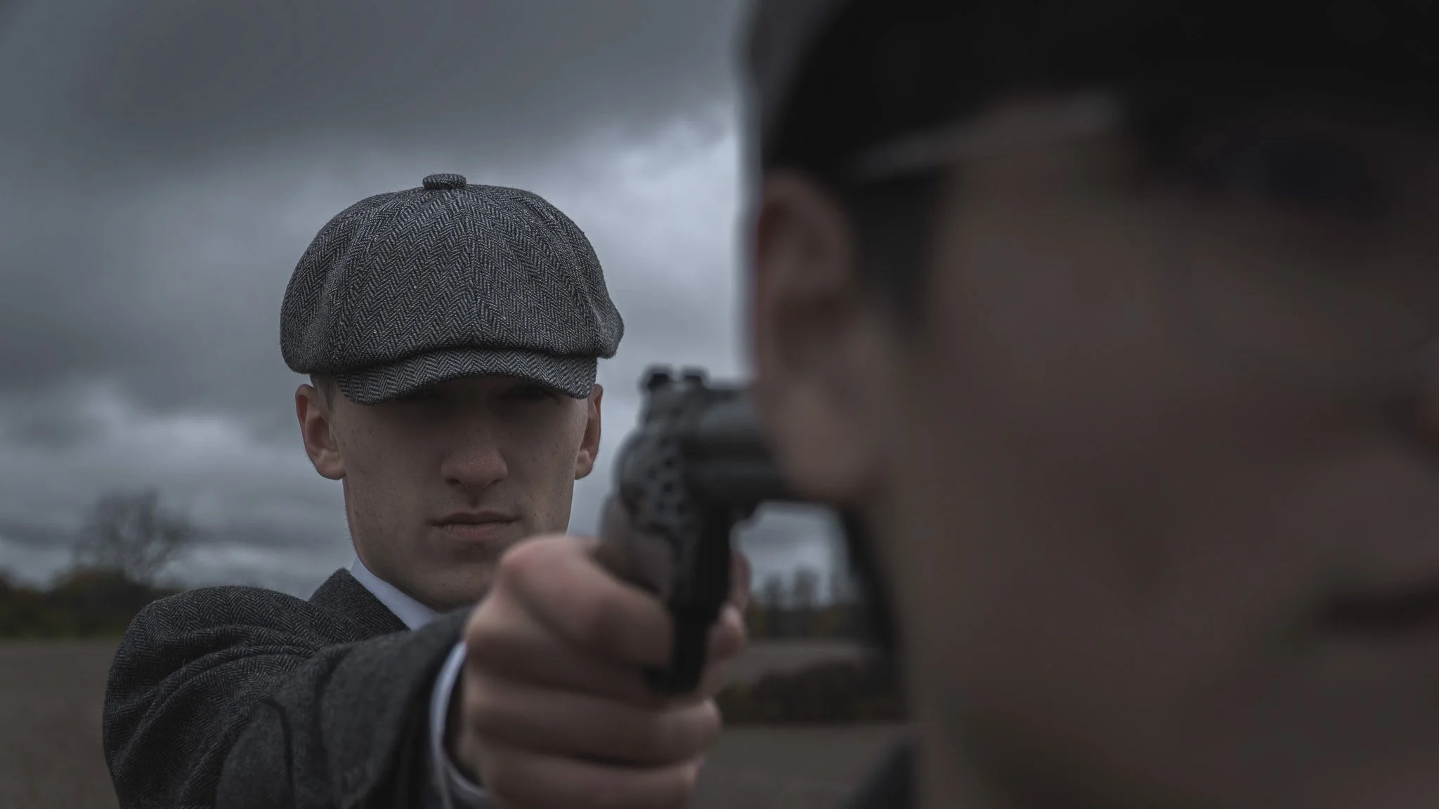 A young man in vintage clothing, including a tweed cap, points a gun at another person whose face is partially visible in the foreground, with cloudy sky in the background.