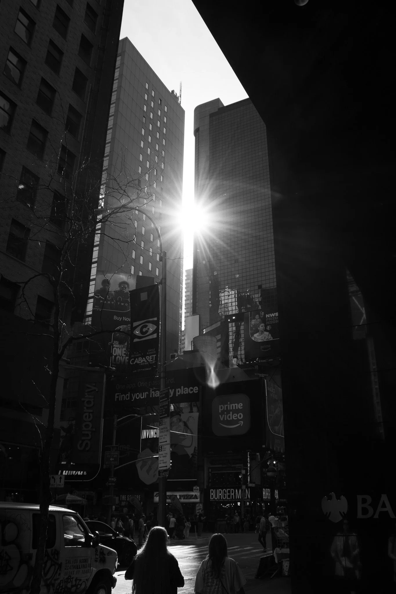 A black-and-white photo of a busy city street with tall buildings, people walking, cars, and various billboards, with the sun shining brightly between the buildings.